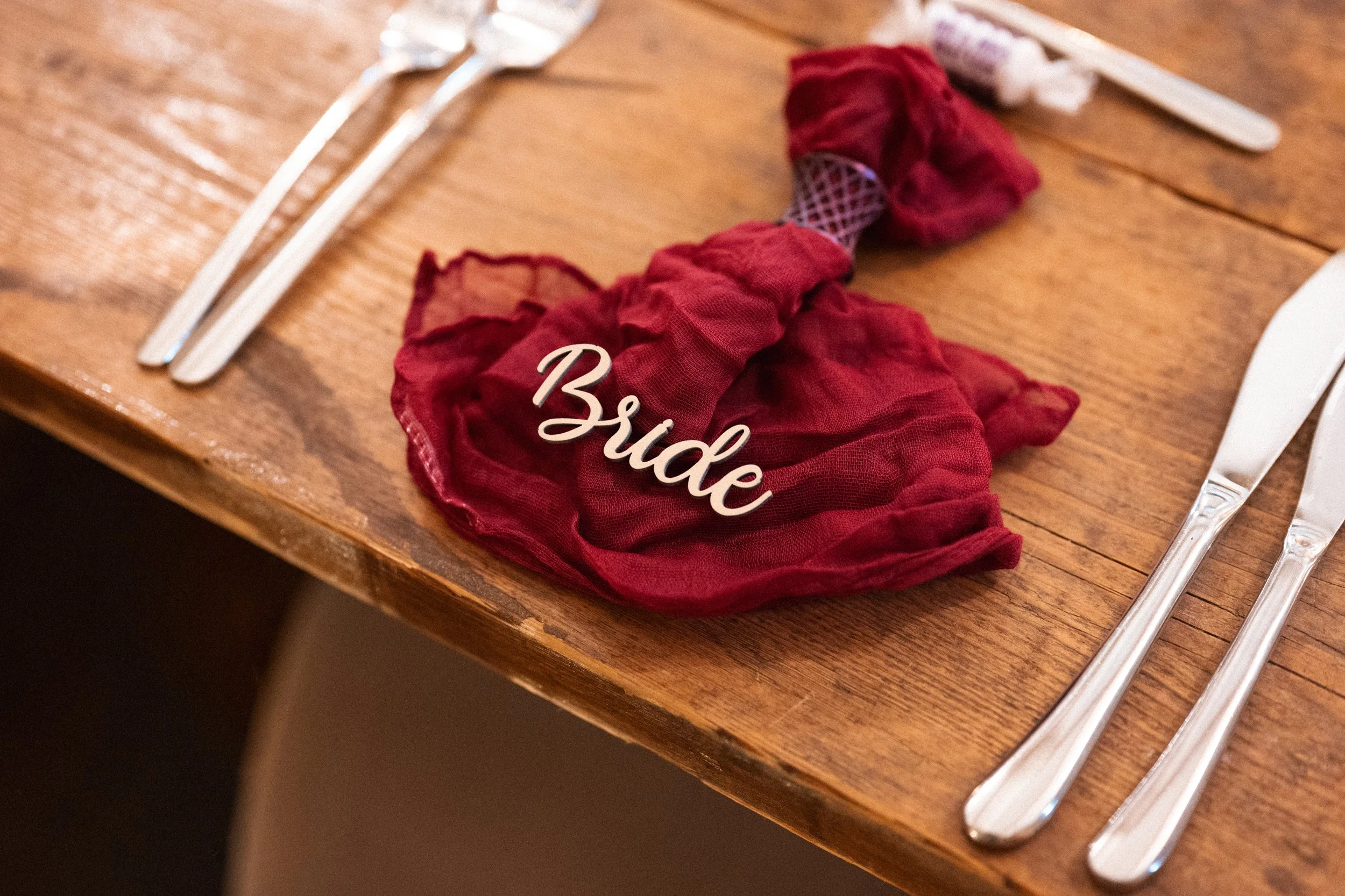 Folded burgundy cloth napkin with the word 'Bride' in white cursive letters, on a wooden table set with silverware.