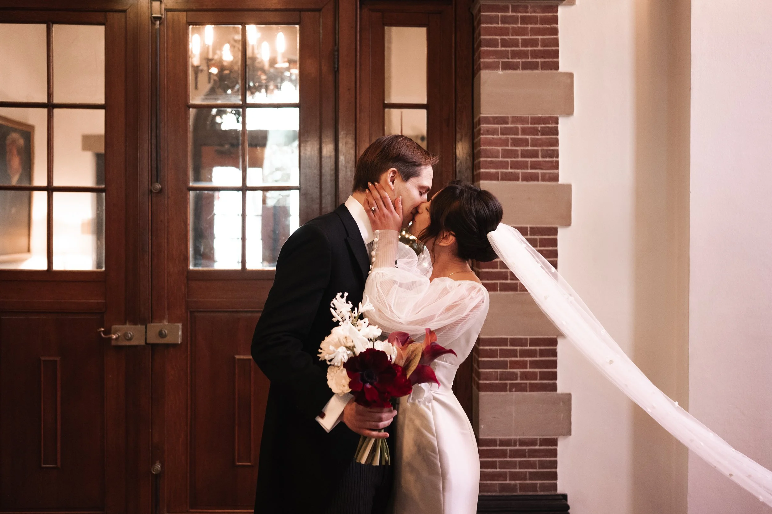 A newlywed couple sharing a kiss indoors. The groom is wearing a black suit and holding a bouquet of white and red flowers. The bride is wearing a white gown with a veil. They are standing in front of wooden doors with glass panes.