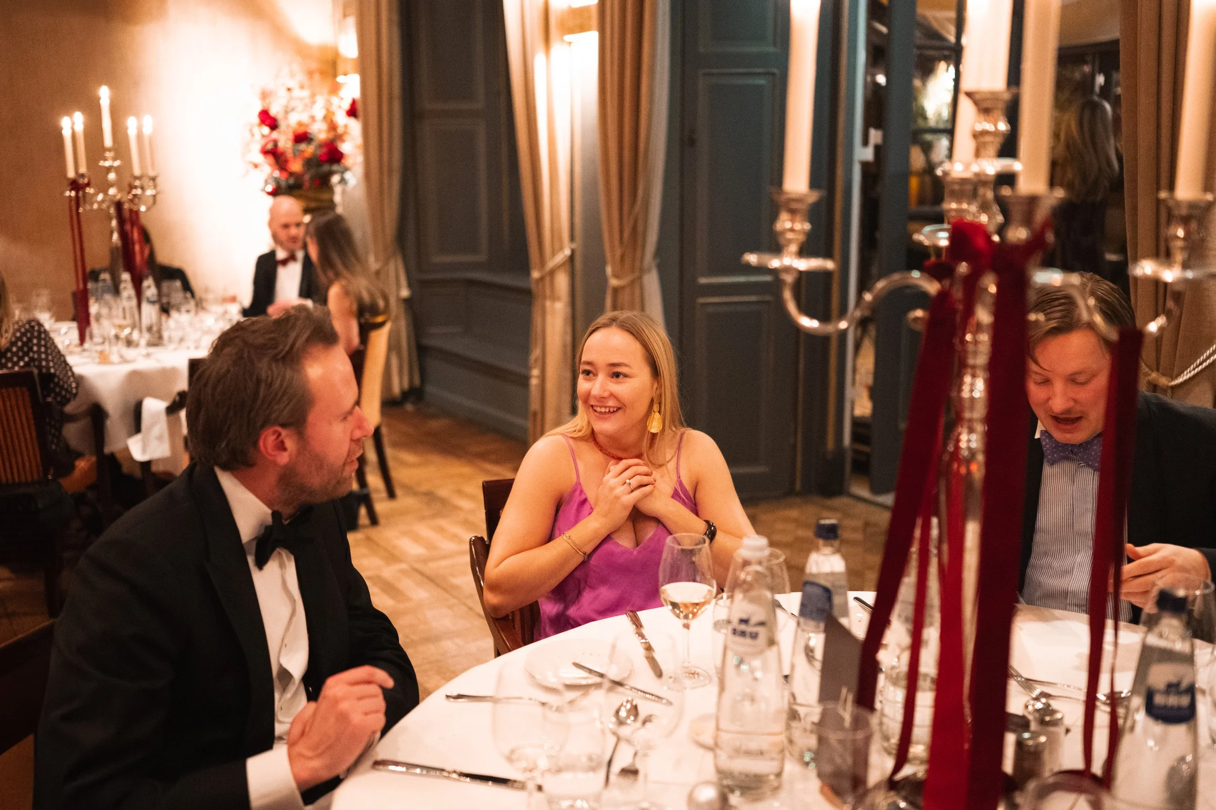 People at a formal dinner party sitting around a table with elegant tableware, a candelabra, and bottles of water, in a decorated, warmly lit room.