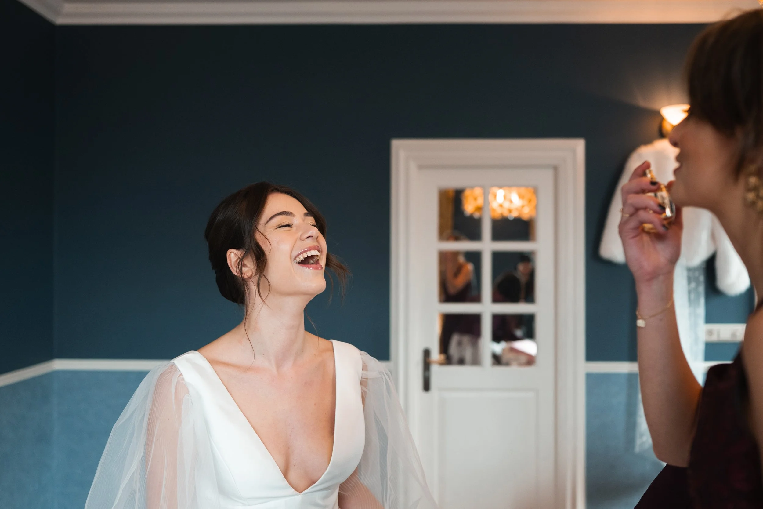 A woman in a white dress laughing and smiling, standing in a room with dark blue walls, while another woman with earrings is holding a perfume bottle near her face.