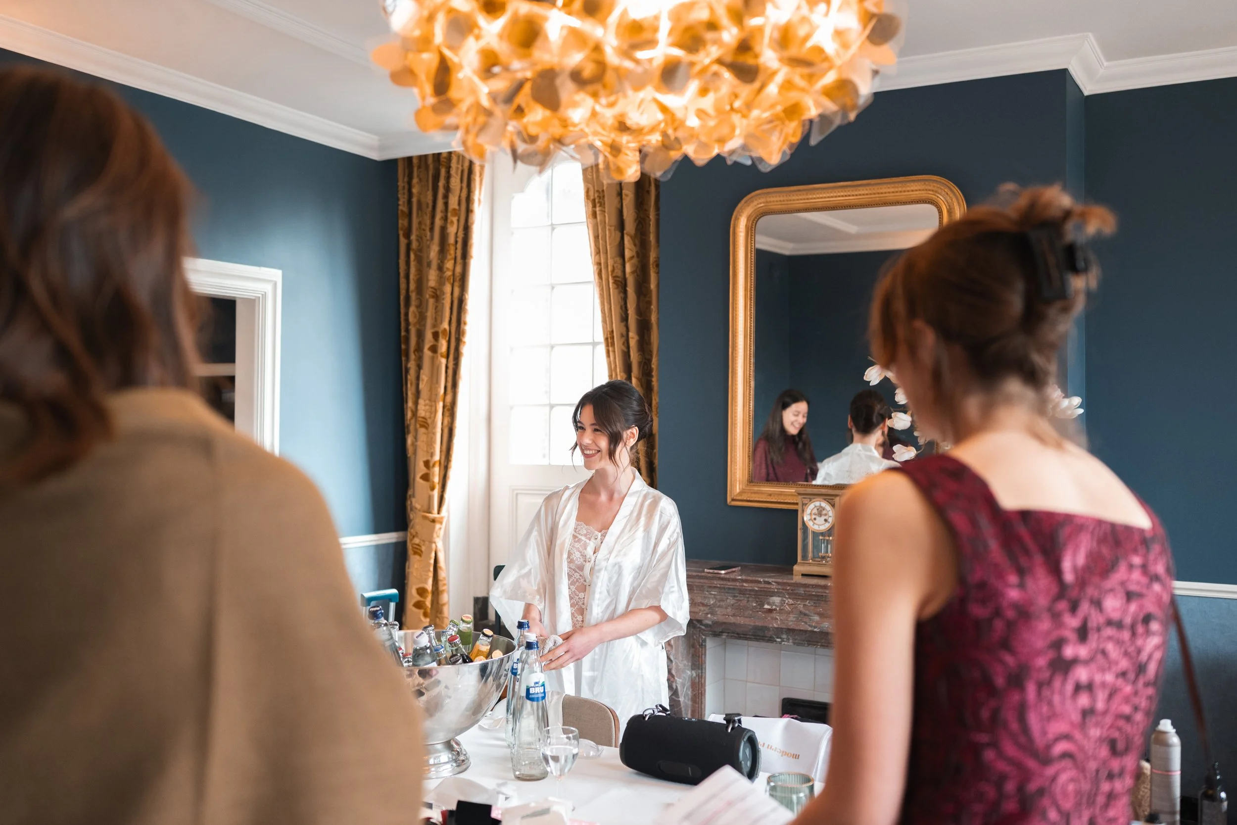 A woman dressed in white smiling while standing near a table with bottles of beverages, in a room with dark blue walls, gold curtains, and a large mirror reflecting her and two other women in elegant attire.