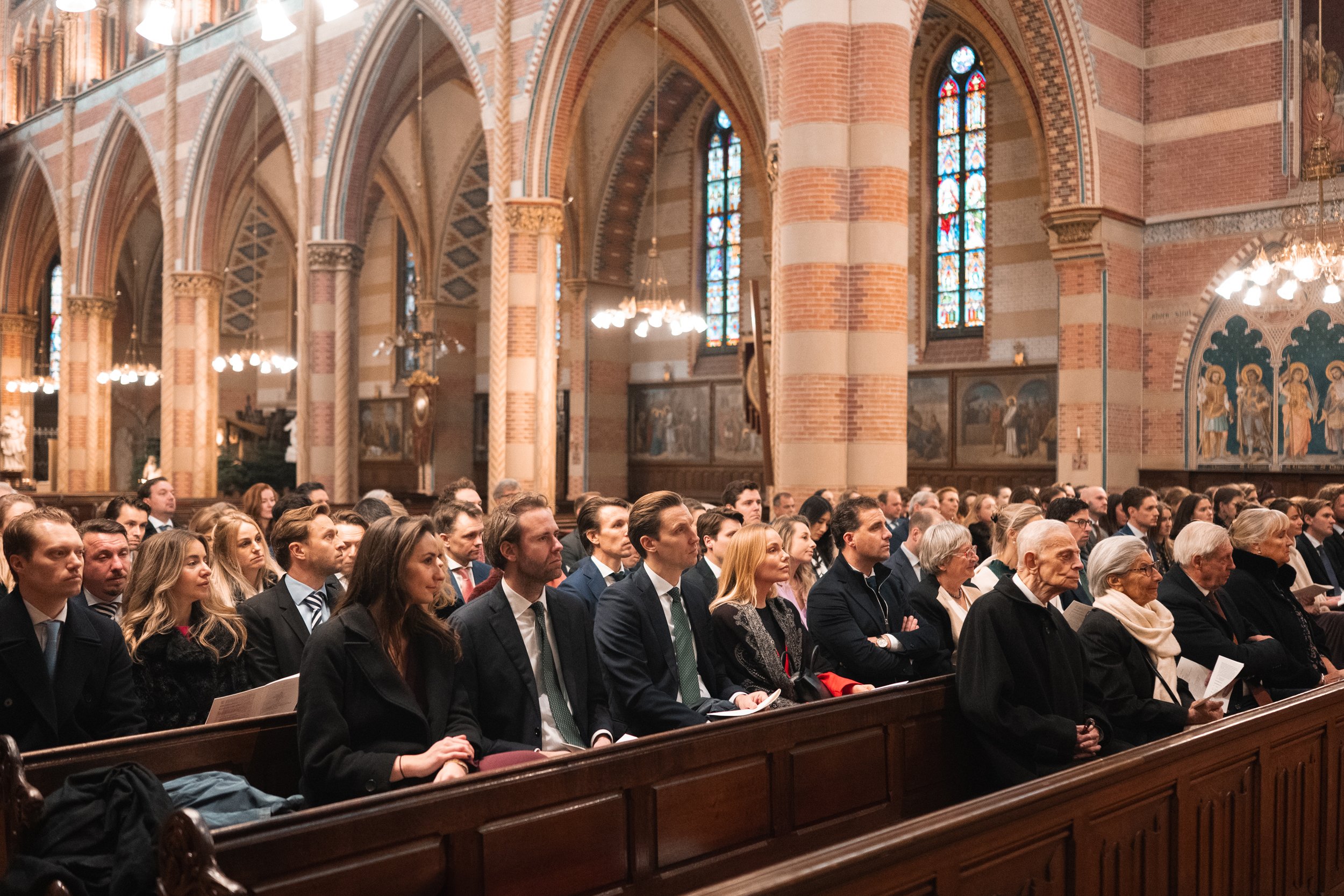 People attending a church service, sitting on wooden pews inside a church with gothic architecture, stained glass windows, and religious artwork.