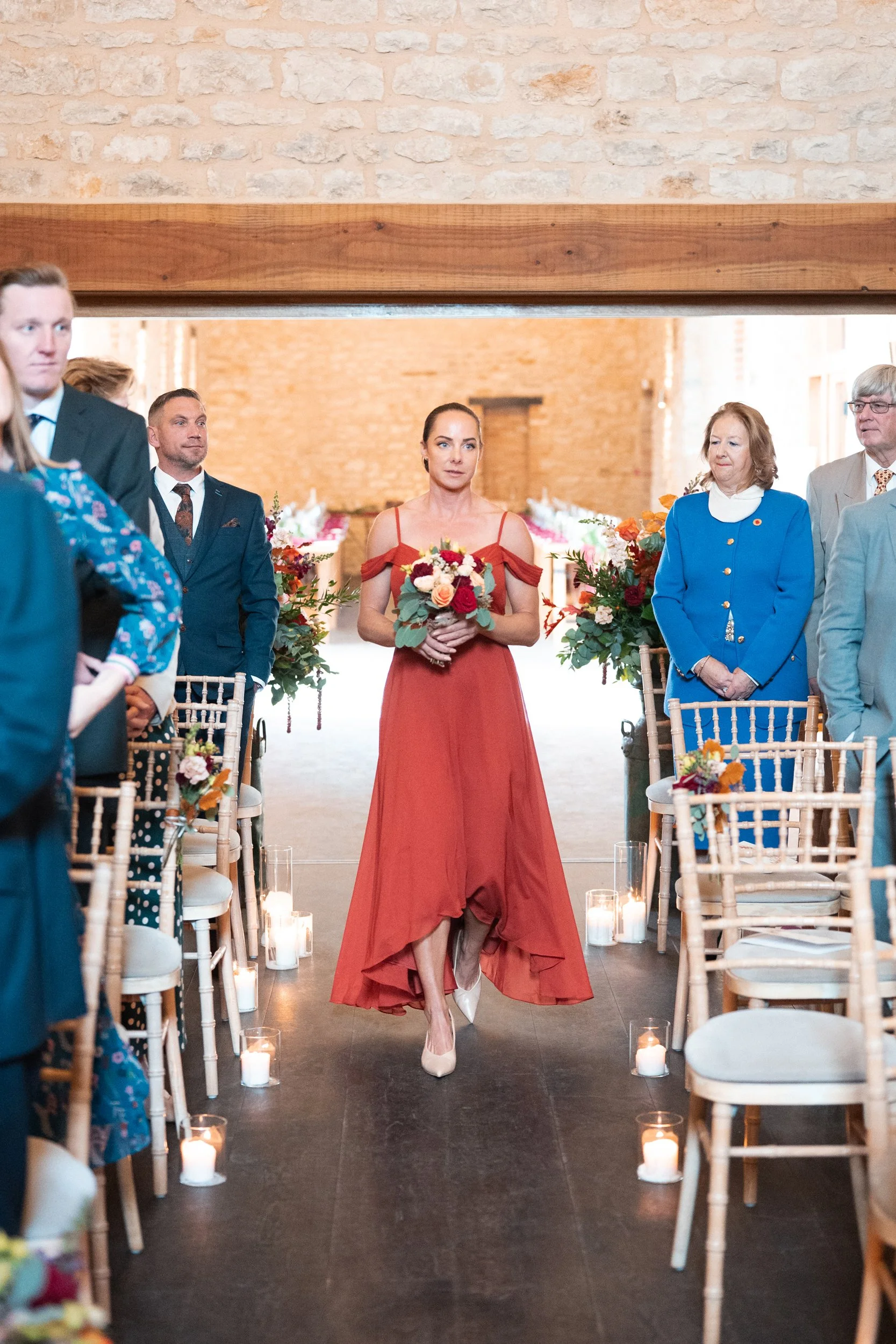 A woman in a red dress walking down the aisle at an indoor wedding, holding a bouquet of flowers, with seated guests on each side and candles on the floor.