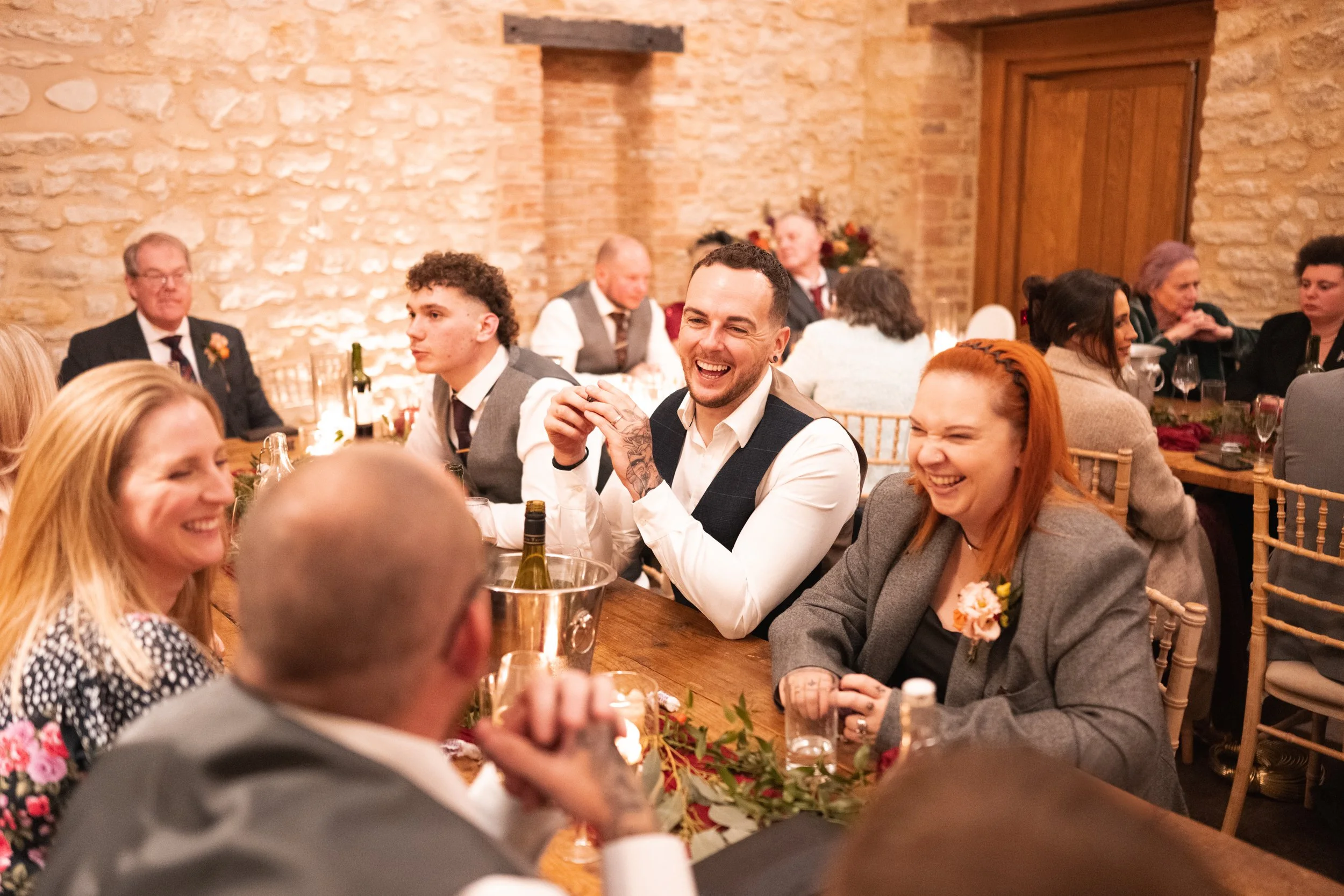 People sitting at a wooden table during a celebration in a cozy venue with brick walls, smiling and laughing, with wine bottles and glasses on the table.