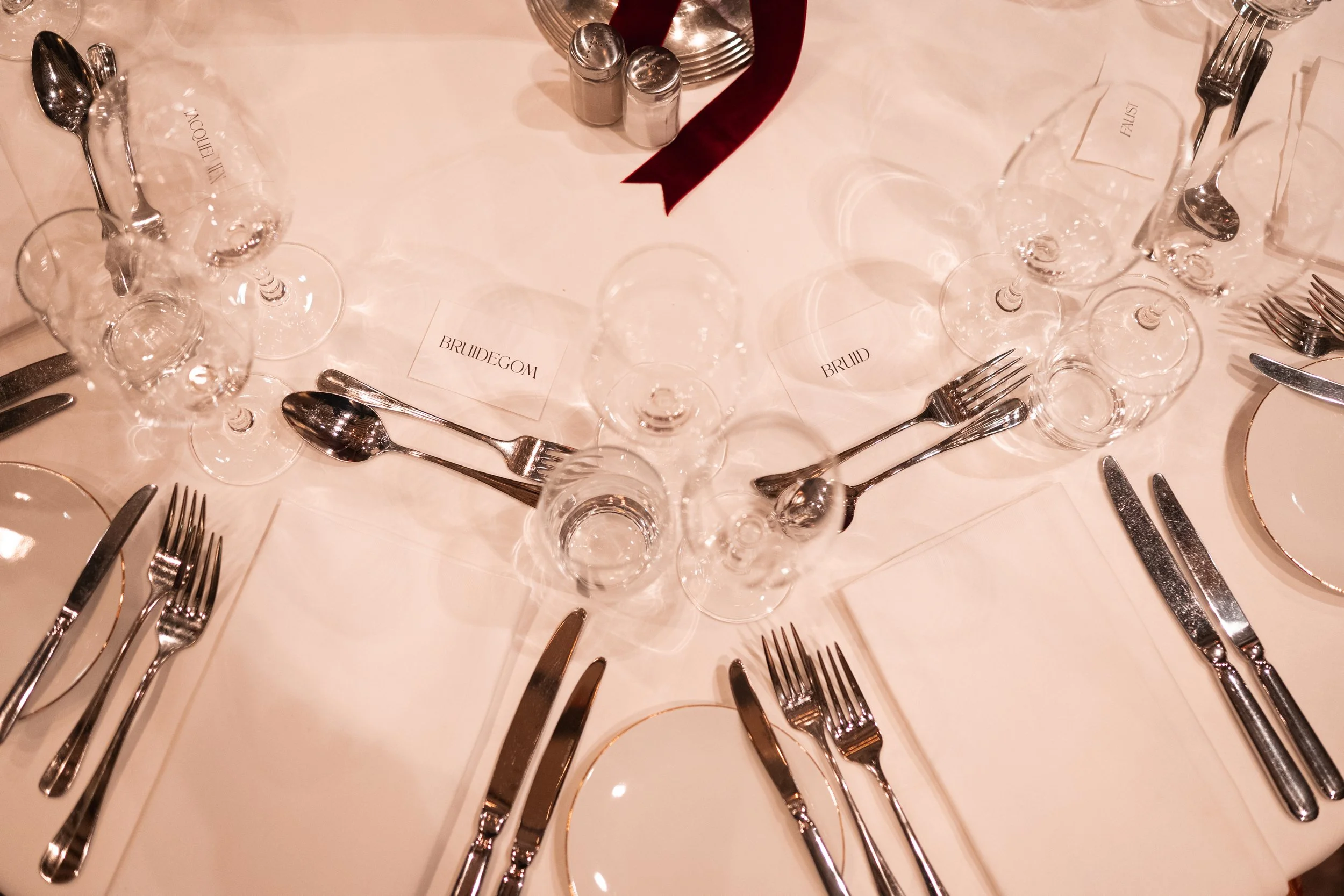 Wedding table setting with multiple glasses, silverware, and name cards labeled 'BRUIDEGOM,' 'BRUID,' and 'BRUIDEGOM,' on a white tablecloth with salt and pepper shakers and a red ribbon.