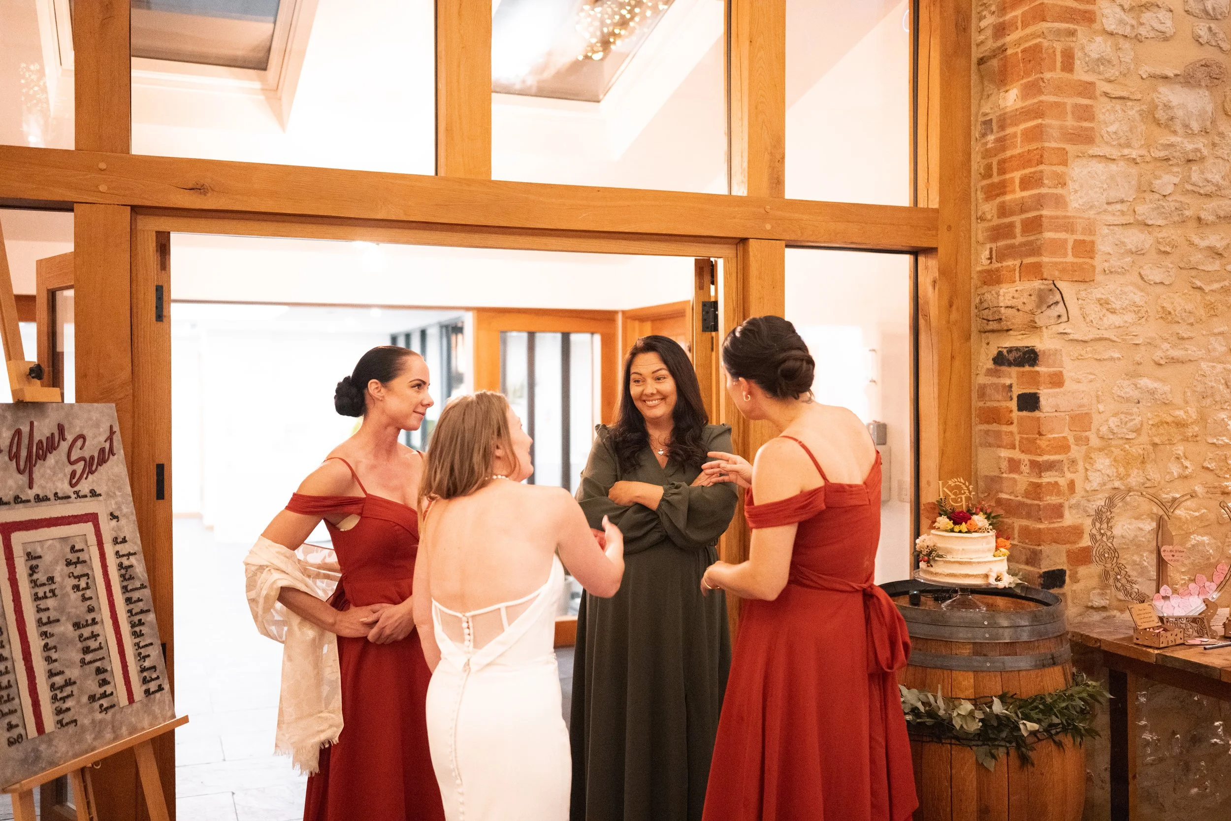 Four women are engaged in conversation at a social gathering, standing near a decorated table with a cake and a decorative wreath.