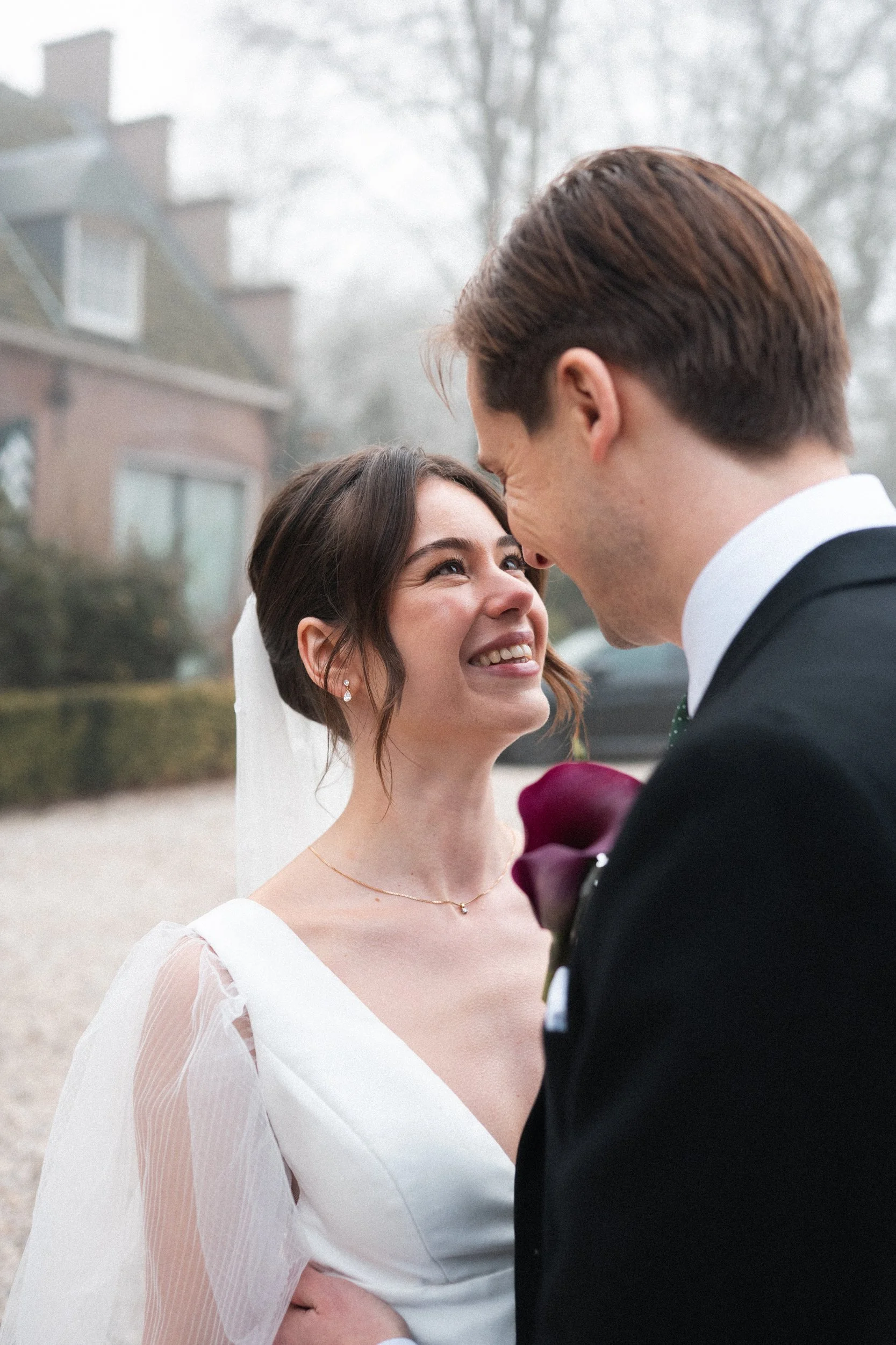 A bride and groom on their wedding day outdoors, gazing into each other's eyes, with a house and trees in the background.