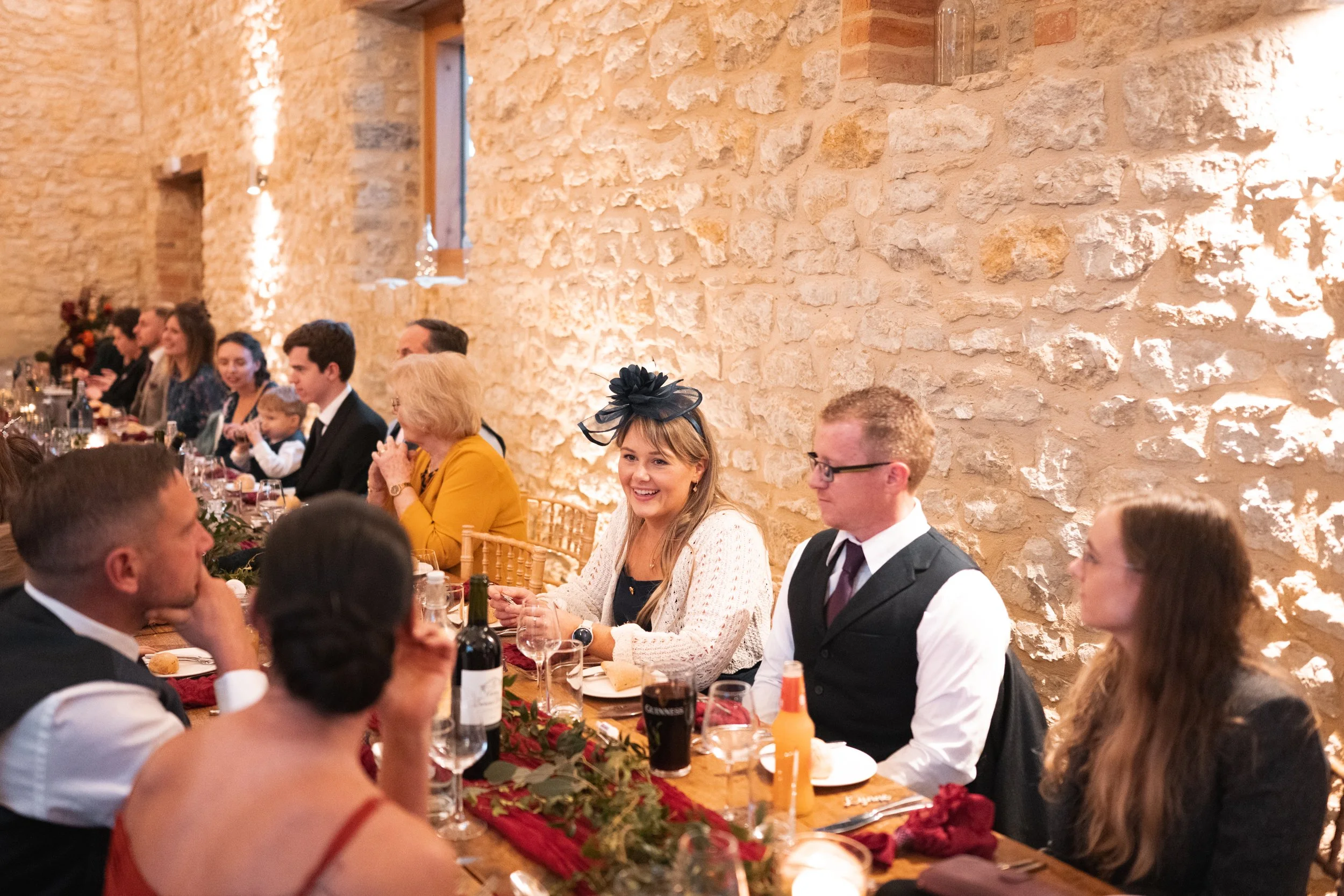 A group of people sitting at a decorated banquet table at a formal celebration, with a woman wearing a black fascinator smiling.