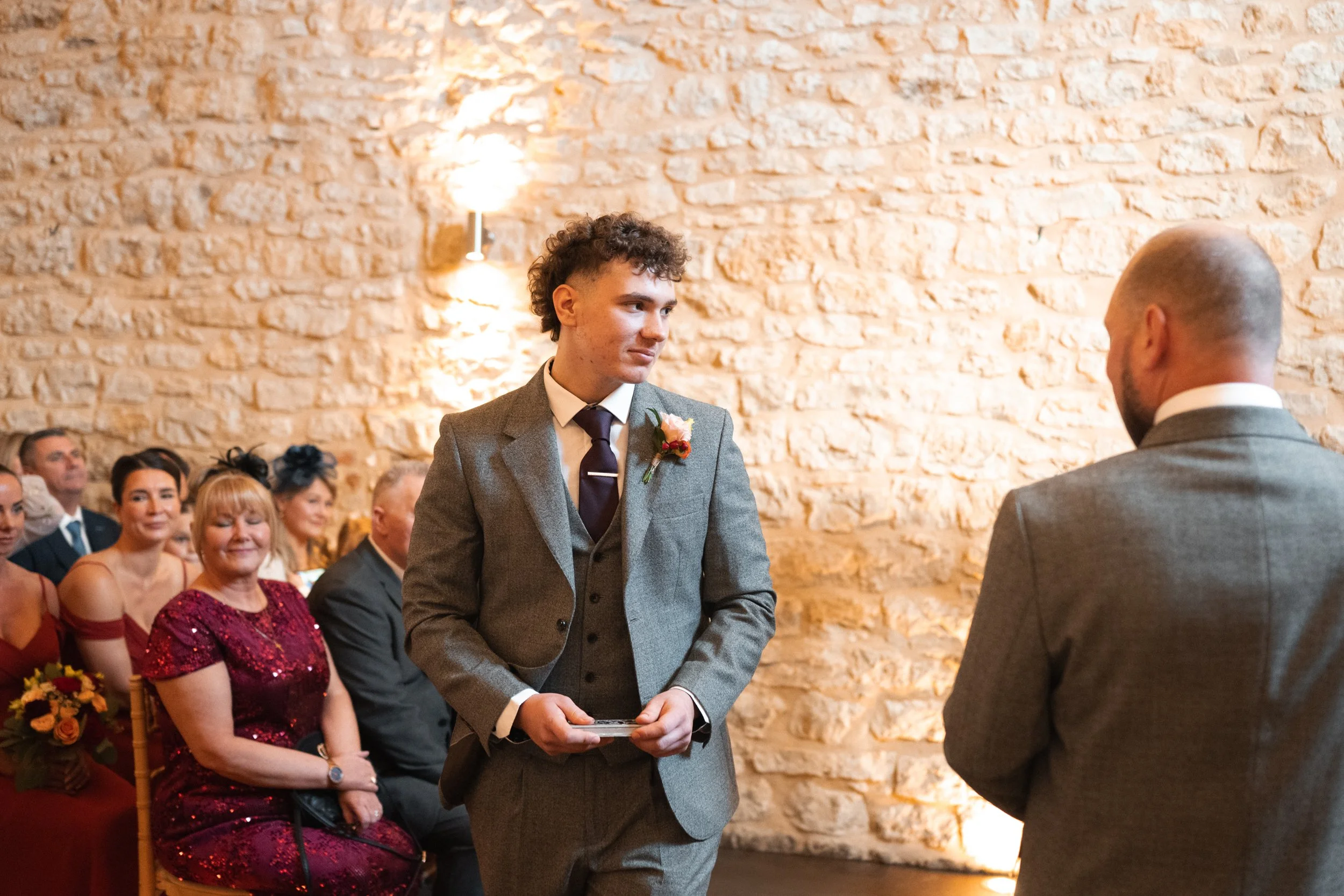 A young man in a gray suit stands during a wedding ceremony, holding a small object, facing another man also in a gray suit. Guests, including women in elegant dresses, are seated on chairs against a brick wall with warm lighting, observing the cerem