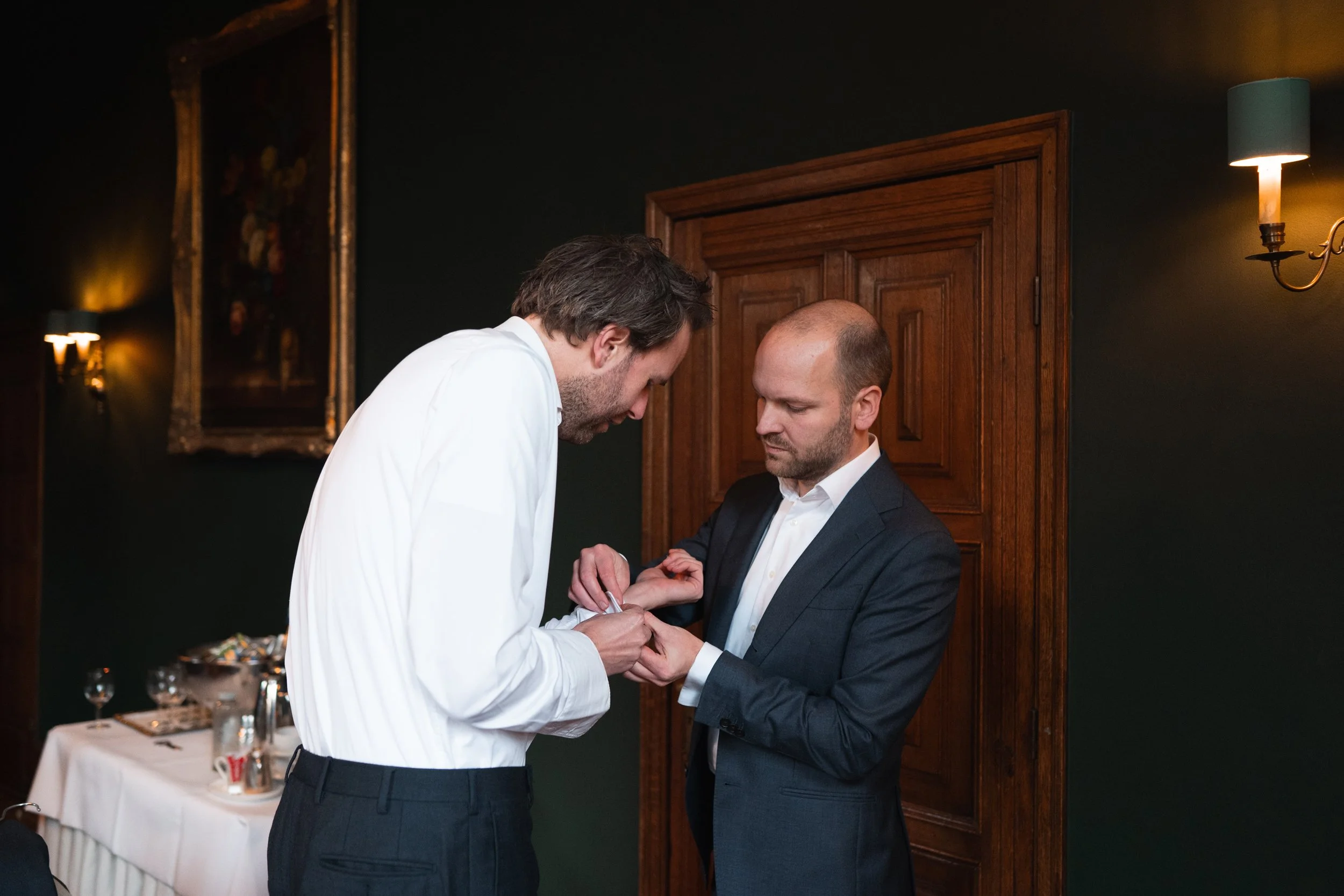 Two men, one in a white shirt and the other in a dark suit, exchanging rings in a dimly lit room with wooden walls and a table with drinks and glasses in the background.