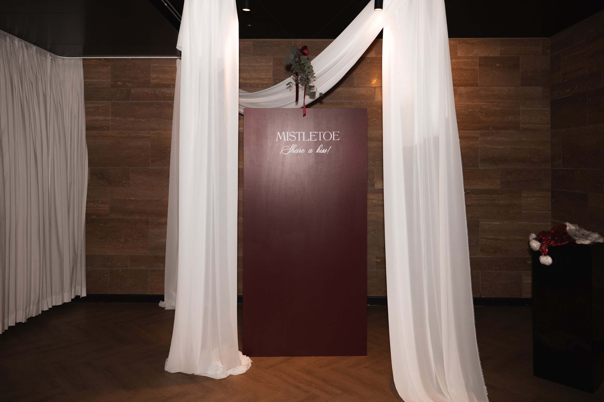 Decorative setup with white curtains, a wooden panel with “MISTLETOE Share a kiss!” written on it, and a small black table with a Christmas hat on top.