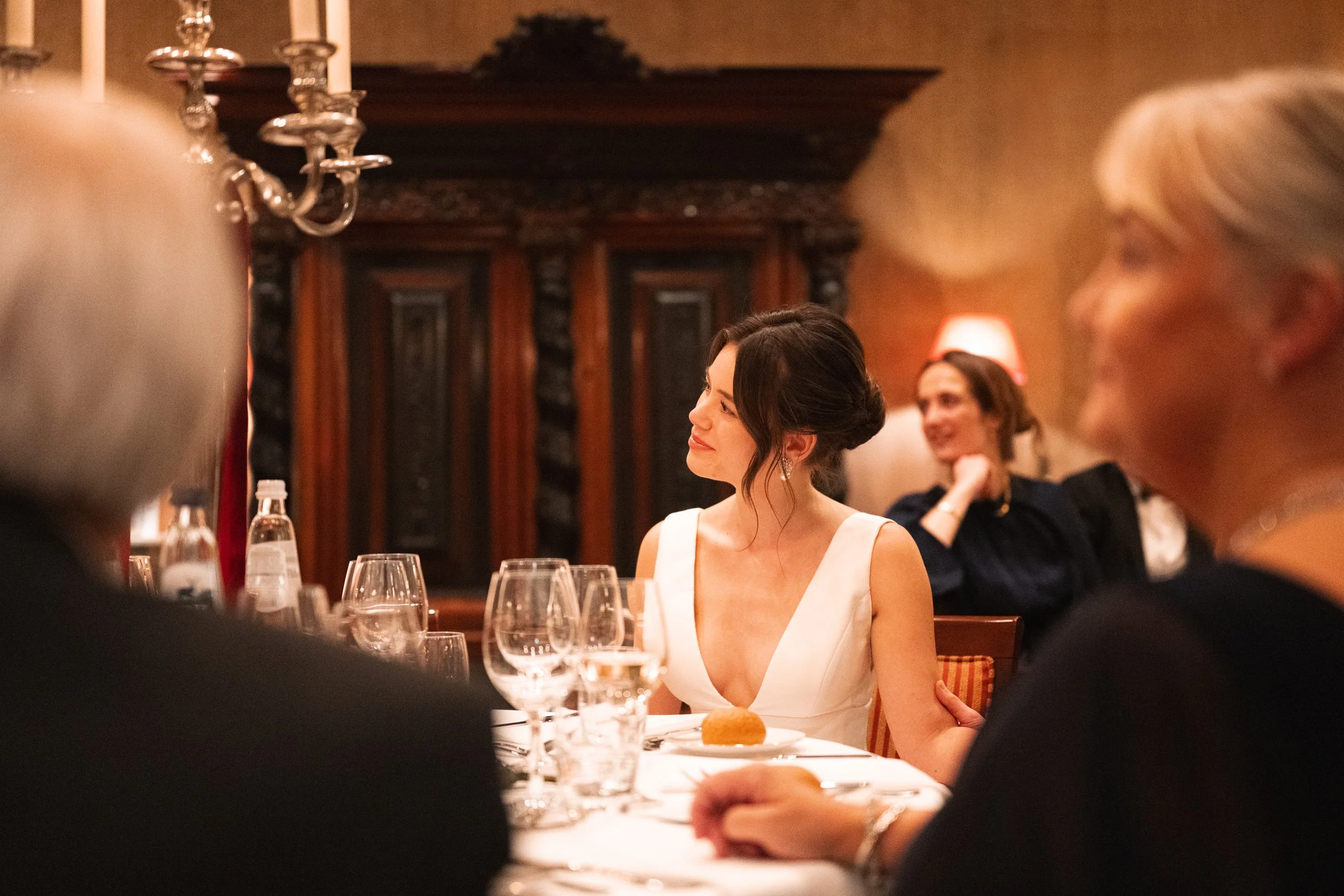 A woman in a white dress sitting at a table during a formal dinner, with others around her, in warm lit ambiance.