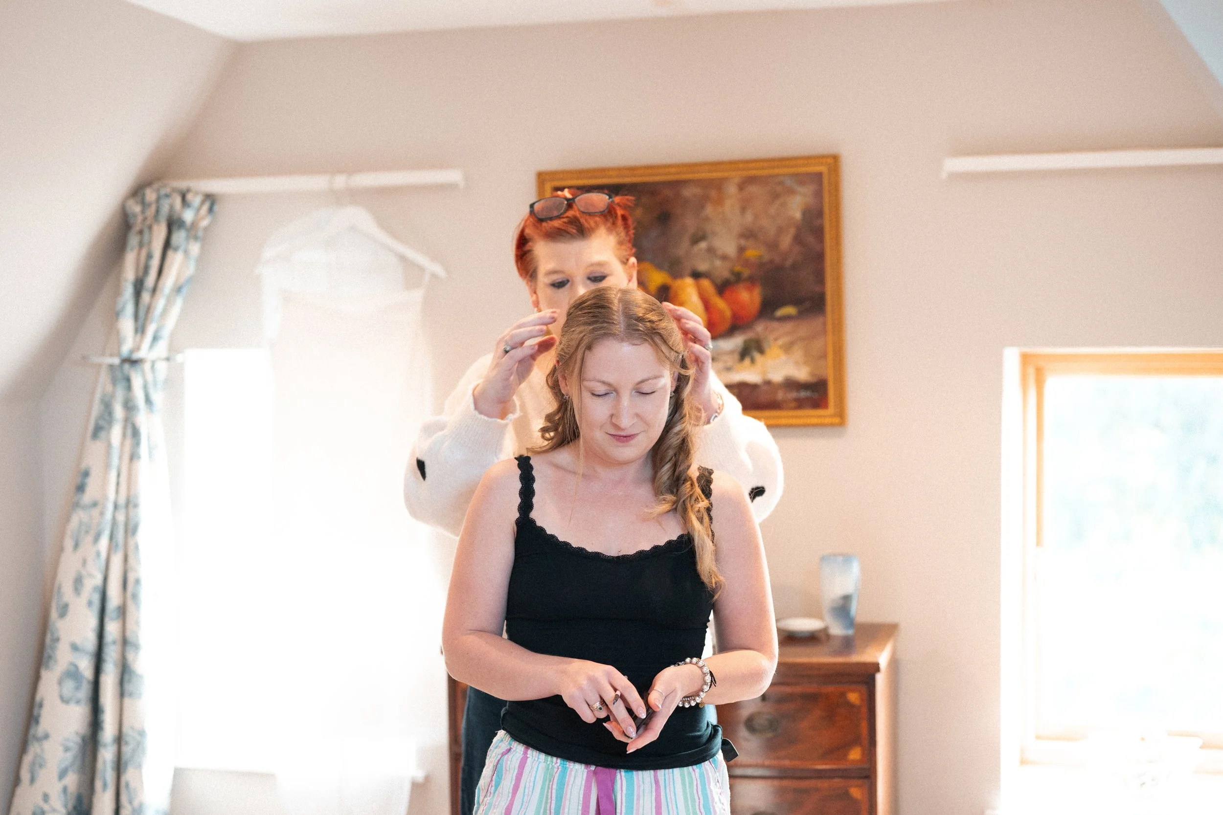A woman in a black top and striped pajama bottoms receives a bridal hair and makeup touch-up from a stylist in a well-lit room.