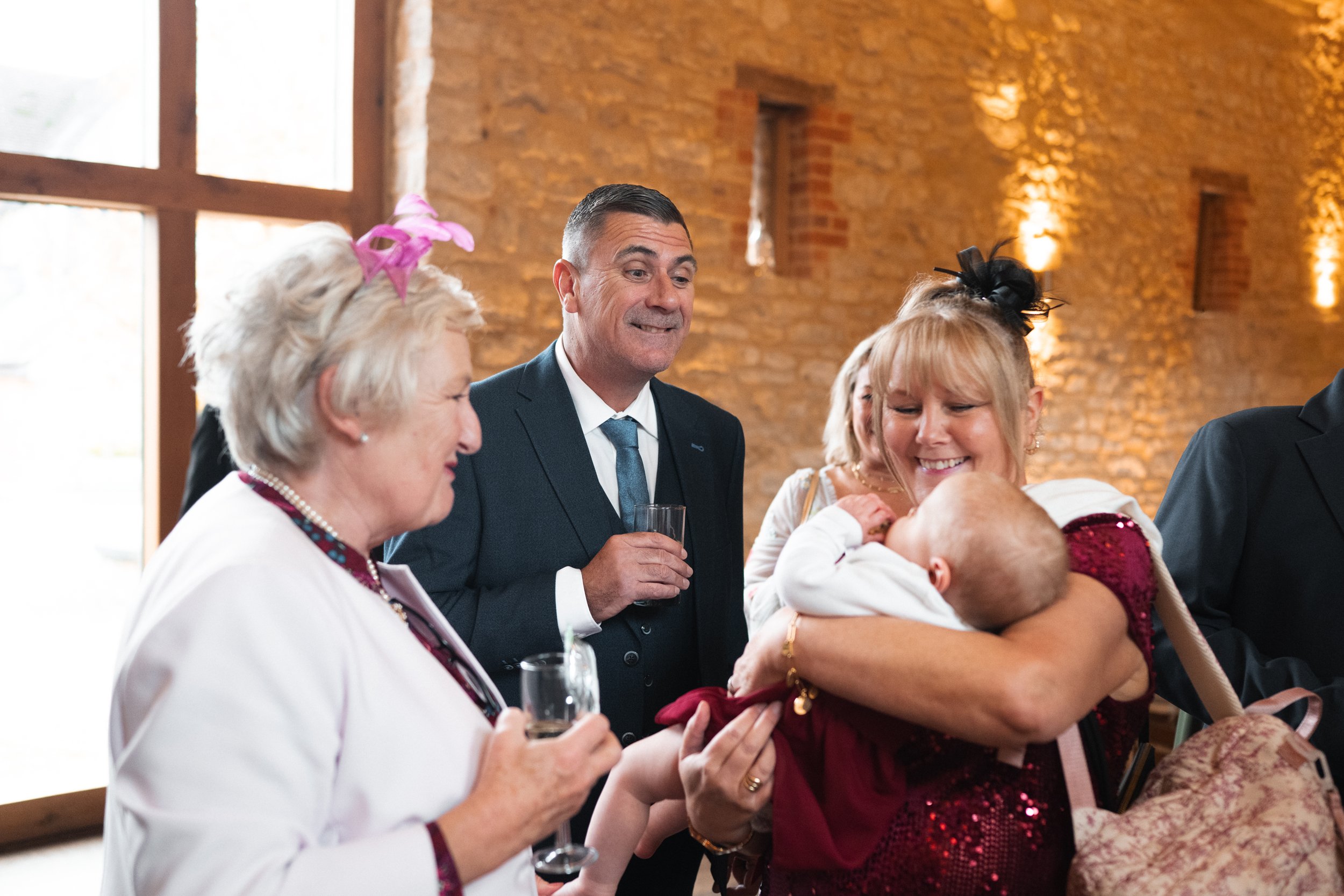 A woman holding a baby and smiling at a social gathering with people in formal attire in a warmly lit room with brick walls.