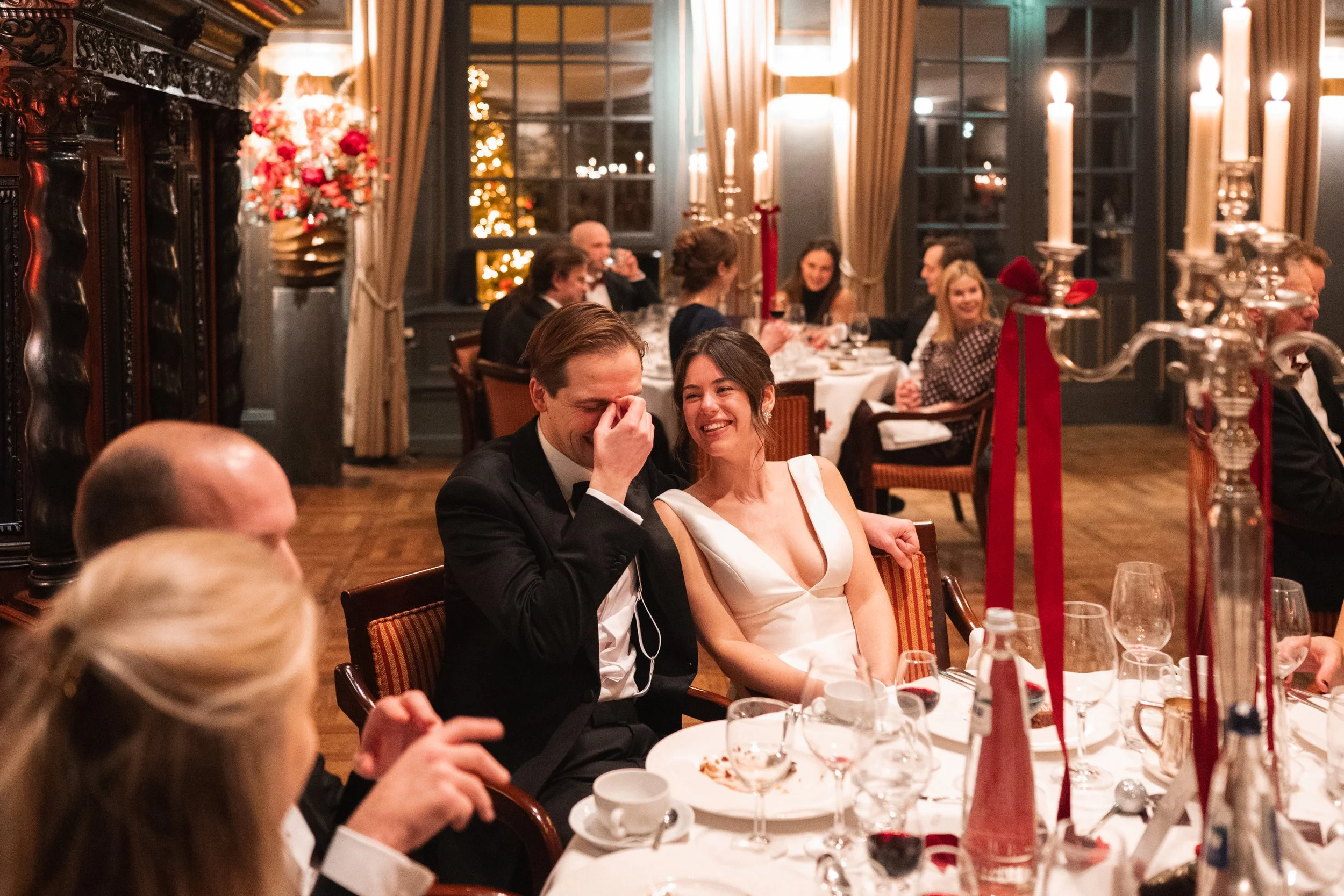A wedding reception with a bride and groom laughing at a decorated table, surrounded by friends and family in a warmly lit, elegant room.