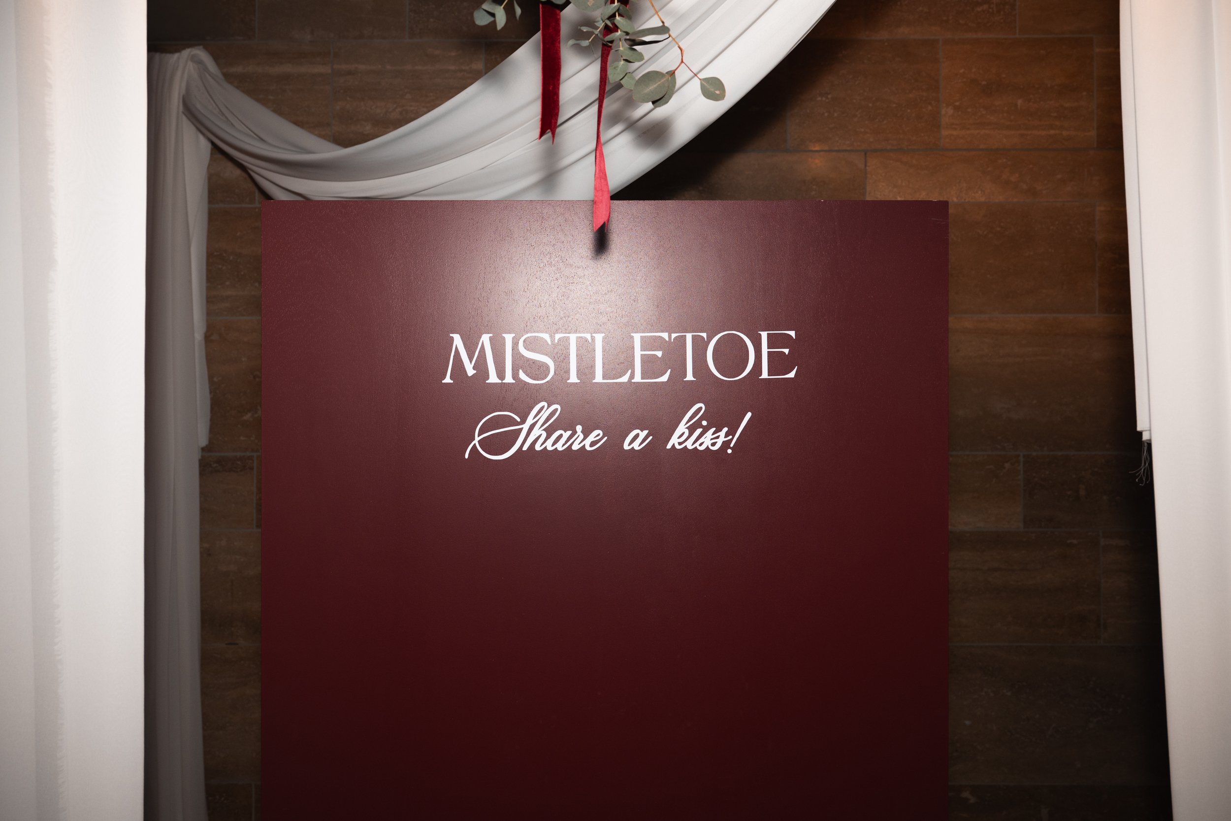 A burgundy sign with white text reading 'MISTLETOE Share a kiss!' hanging from a red ribbon near white curtains and eucalyptus leaves against a wooden wall.