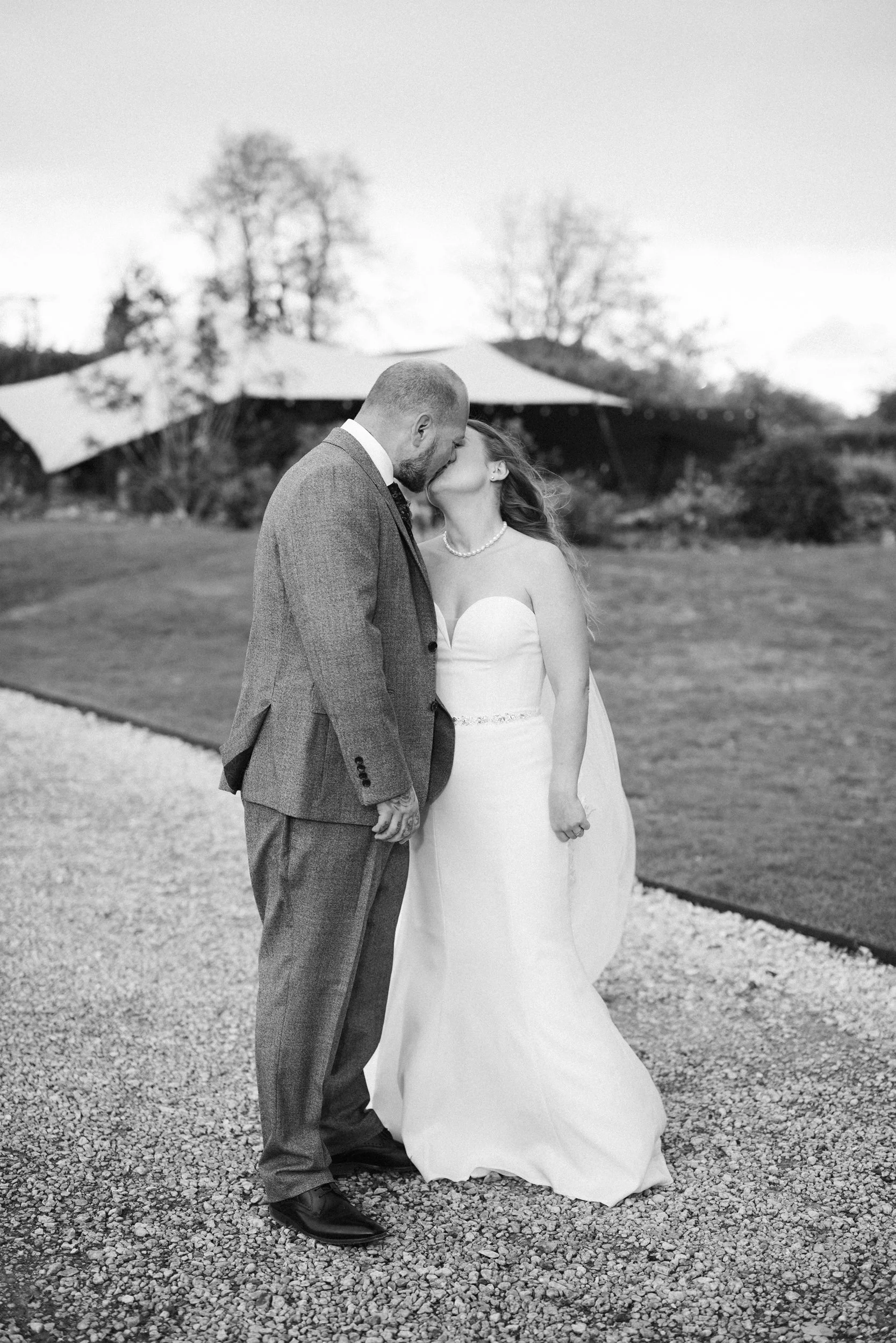 A black and white photo of a couple kissing outdoors. The woman is wearing a strapless wedding dress and the man is wearing a suit. They are standing on a gravel path with trees and a large canopy in the background.