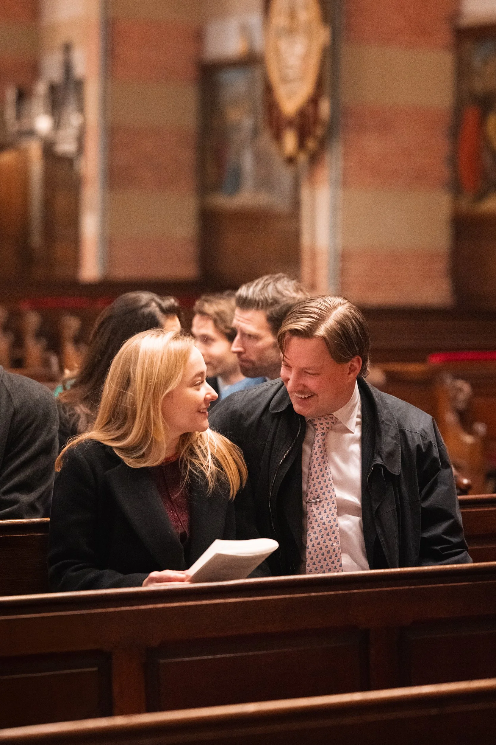 Two people sitting in church pews, engaged in conversation. The woman has long blonde hair and is holding papers; the man has short light brown hair and is wearing a jacket and tie. They are smiling and looking at each other.
