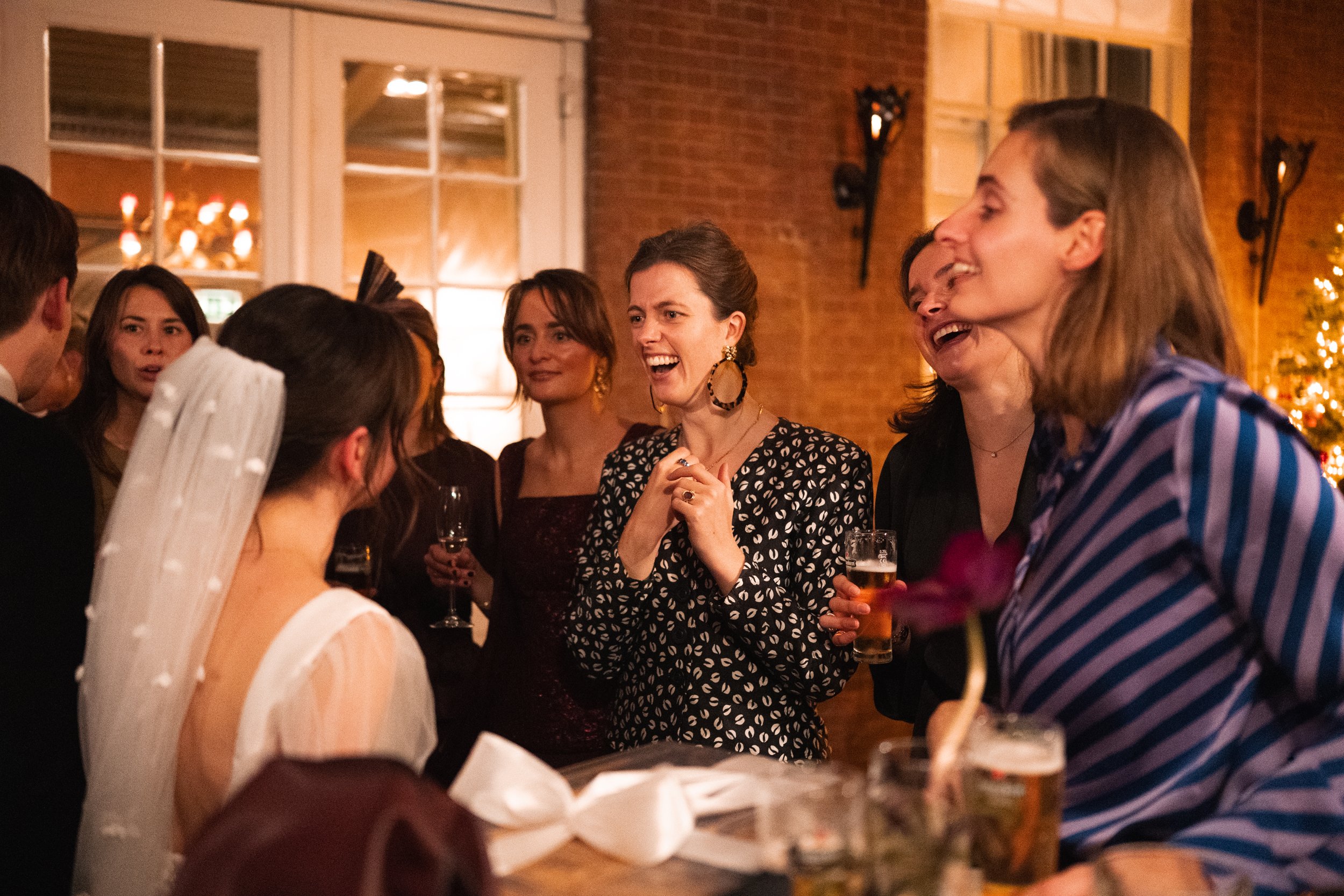 Group of people at a wedding reception, smiling and talking with the bride, wearing a white veil, and other guests holding drinks and enjoying conversation.