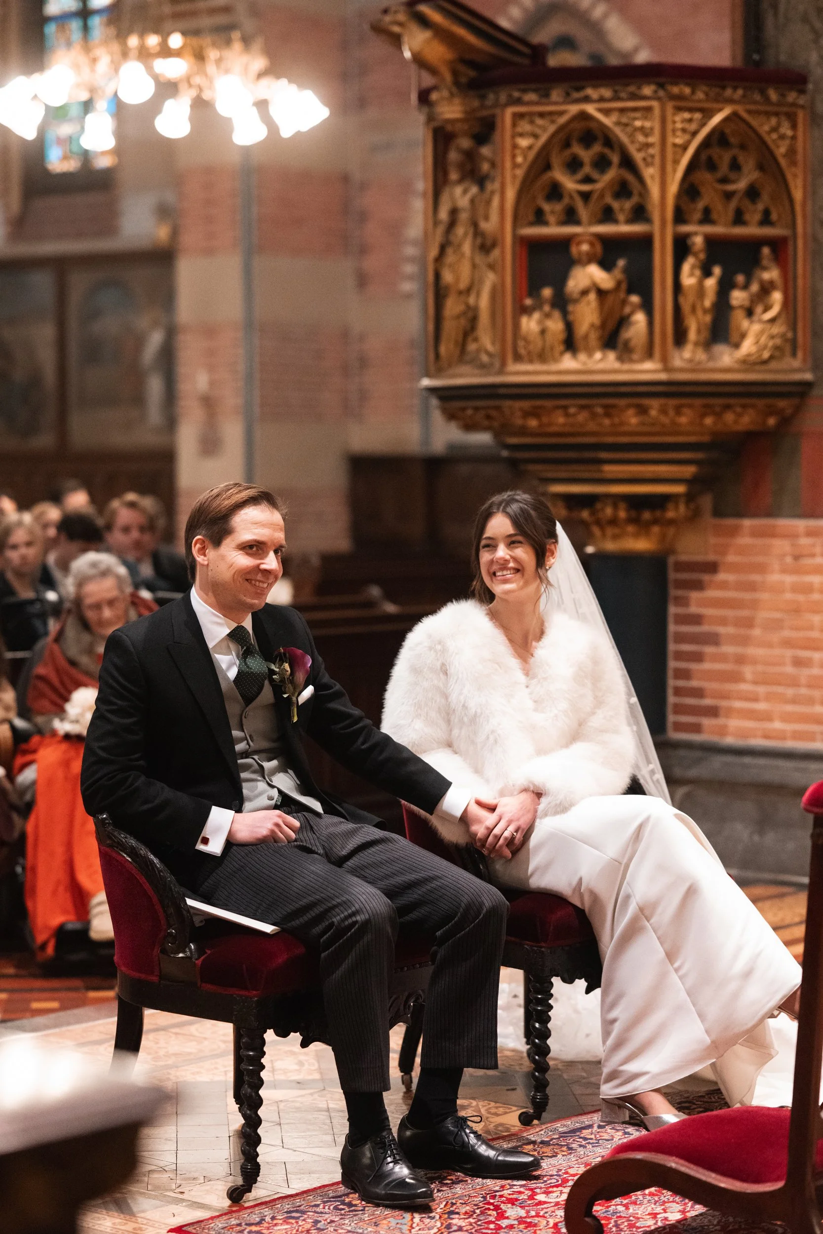 A bride and groom sitting together during a wedding ceremony in a church, holding hands and smiling, with guests seated in the background.
