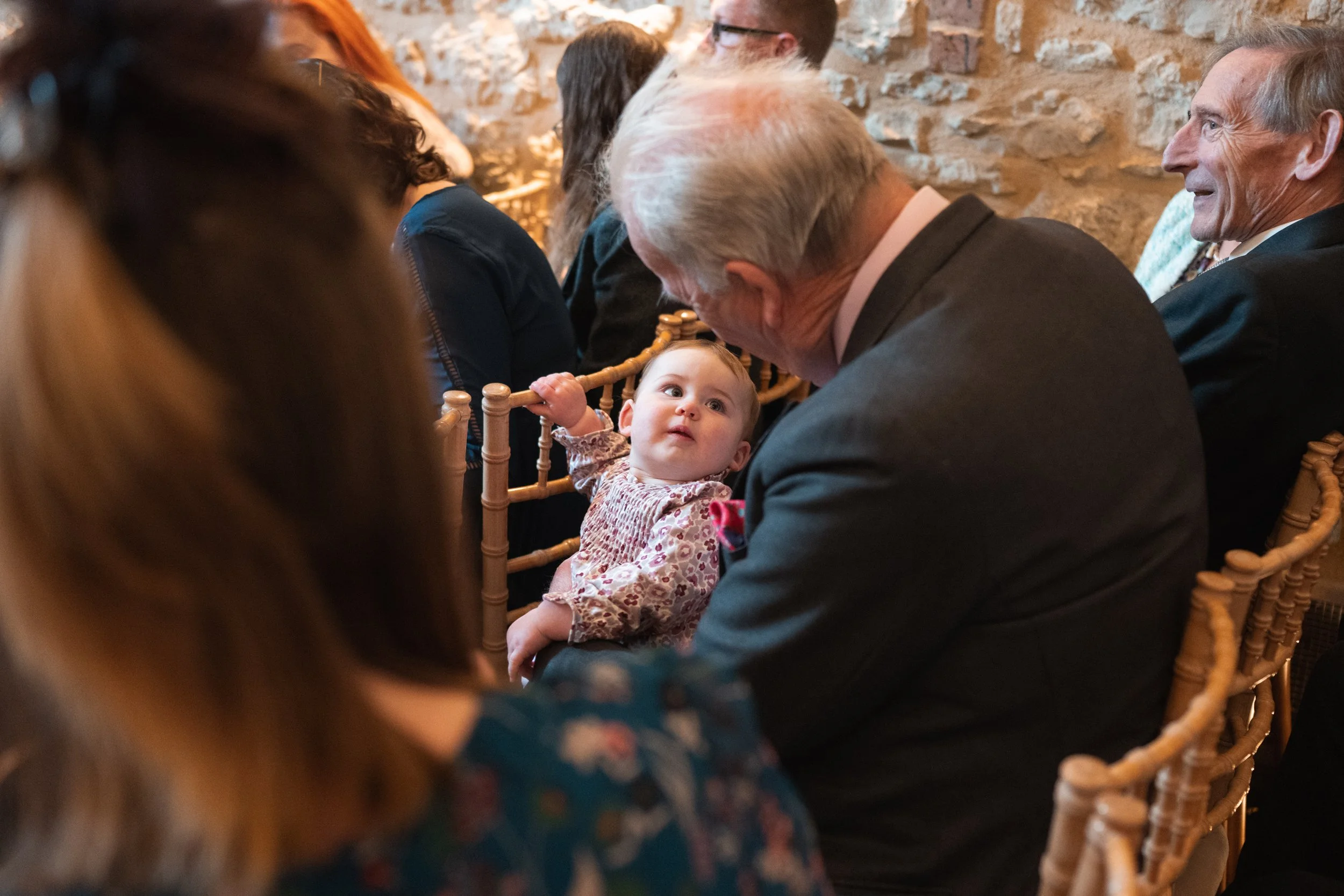 An elderly man holding a baby girl, who is looking up at him, at a formal event with other seated guests in the background.