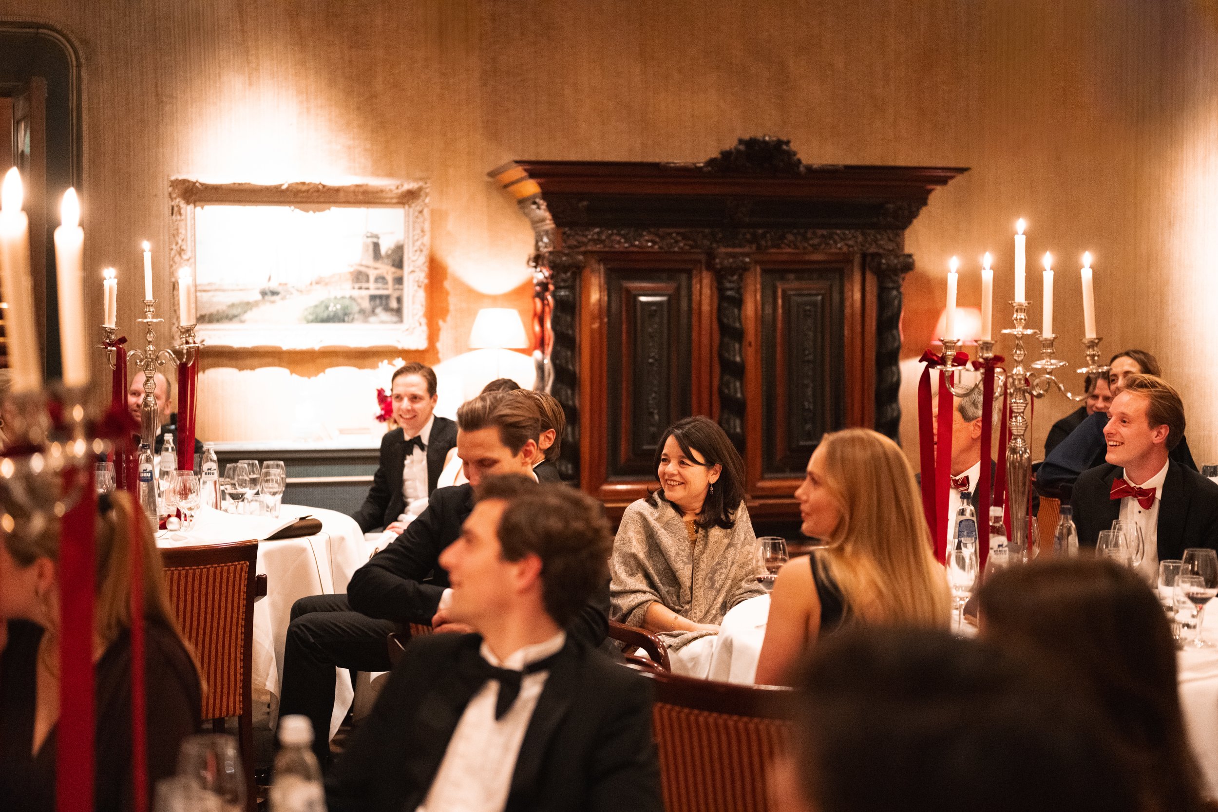 People dressed in formal attire, seated at round tables with white tablecloths and glassware, smiling and listening, in an elegant warmly lit dining room with gold-colored walls, candelabra, and ornate dark wood furniture.