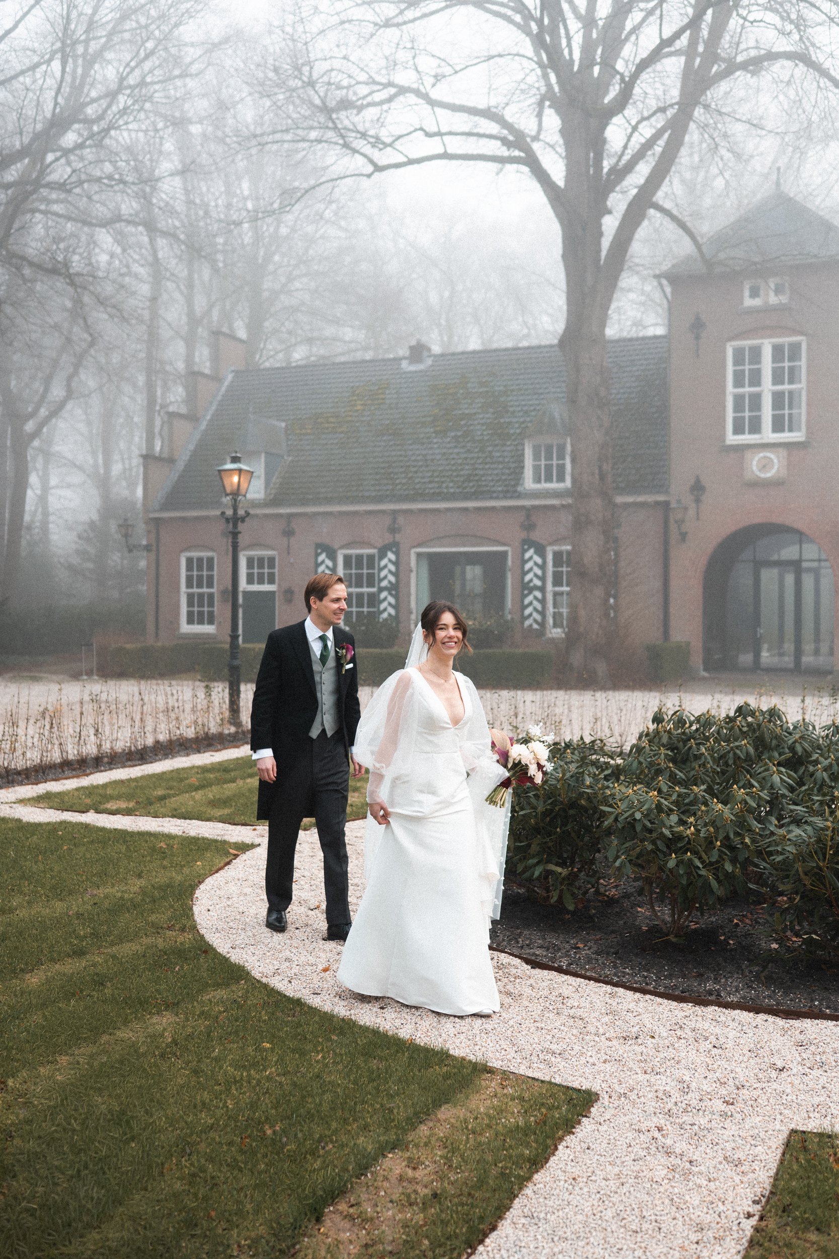 A wedding couple walking along a curved gravel path in a garden, with a foggy house and leafless trees in the background, during daytime.