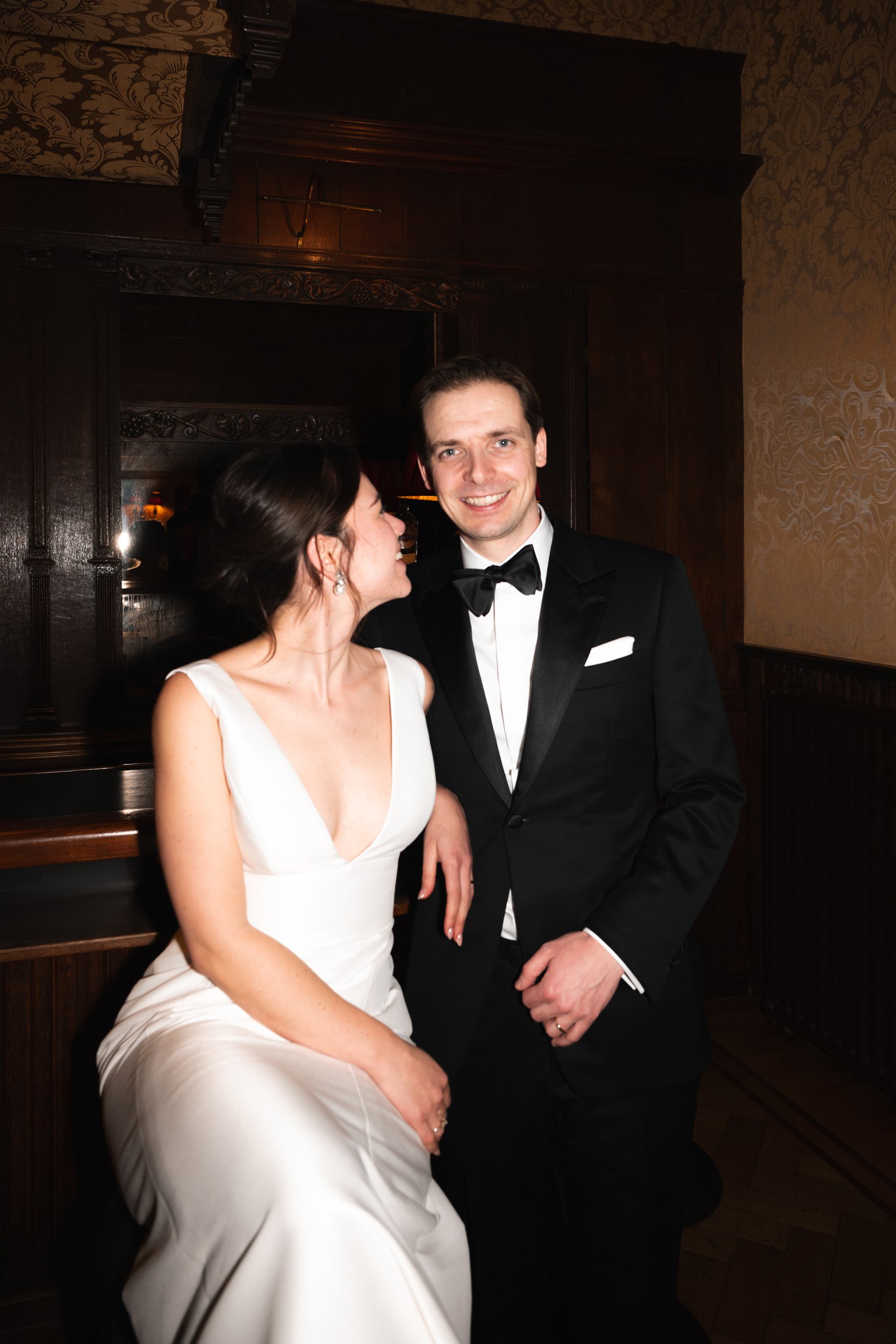 A couple dressed in formal wedding attire, with the woman in a white gown and the man in a black tuxedo with bow tie, posing together in an elegant indoor setting.