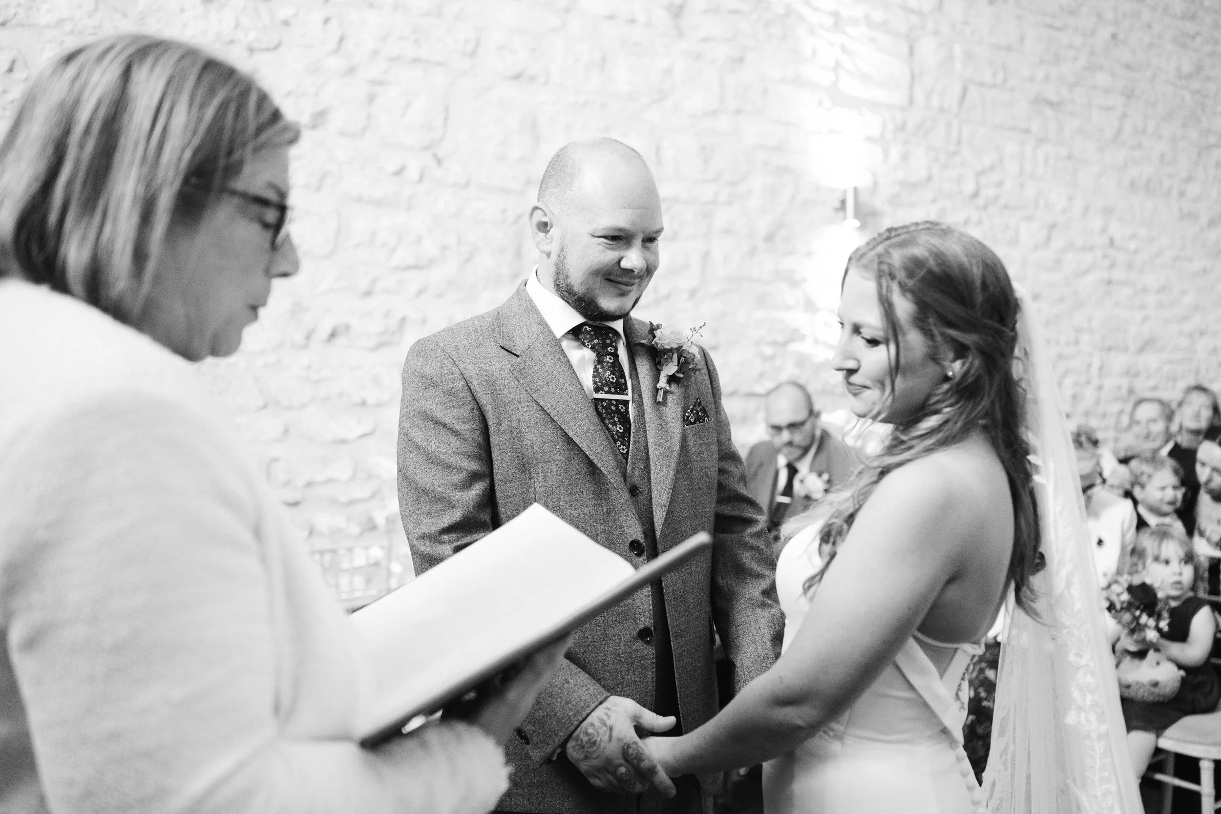 A black and white photo of a wedding ceremony with the bride and groom holding hands, standing before an officiant who is reading from a book, in front of guests seated against a stone wall.
