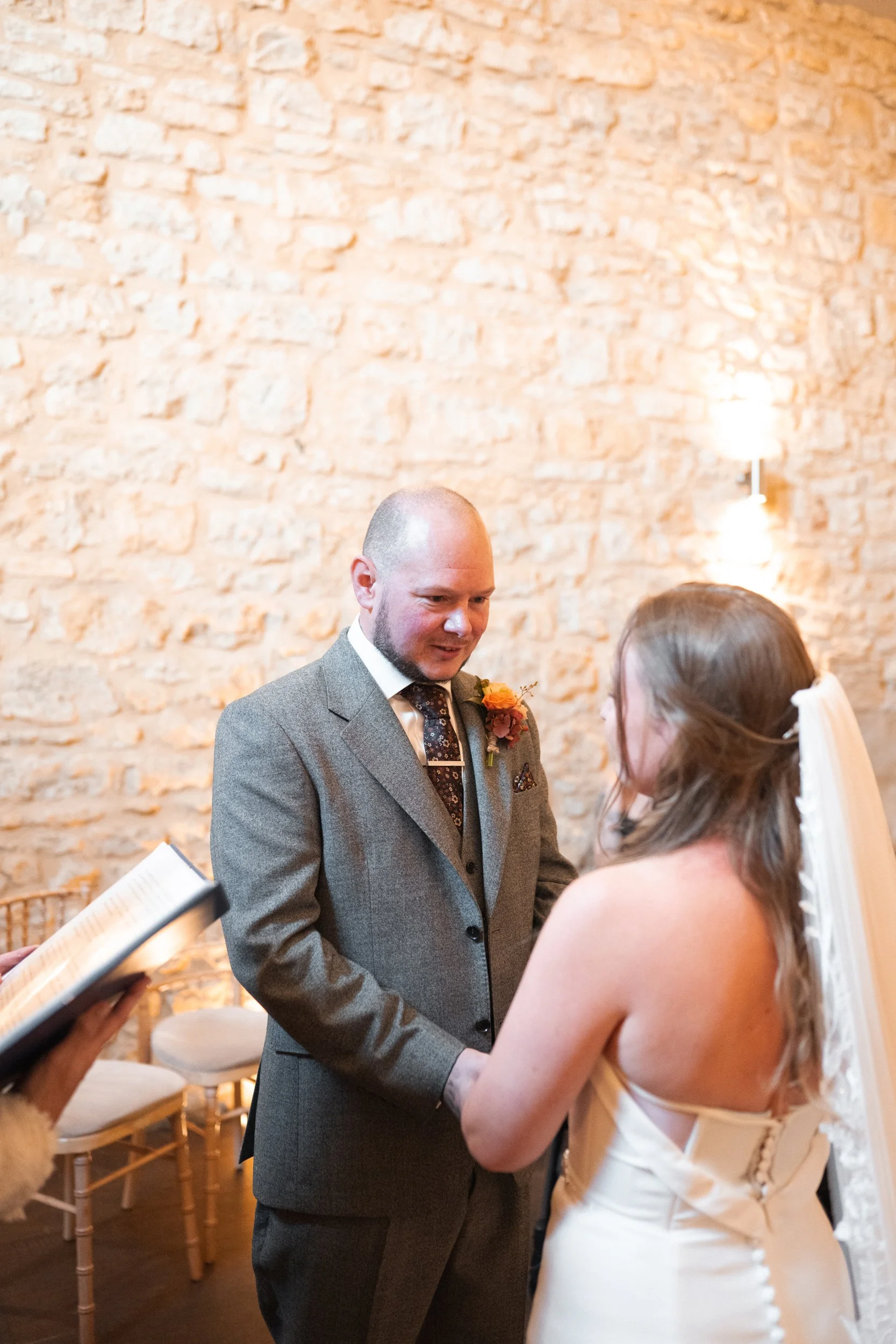 A groom and bride during a wedding ceremony, holding hands and exchanging vows, with a wedding officiant holding a book nearby. The setting has a rustic brick wall background with soft lighting.