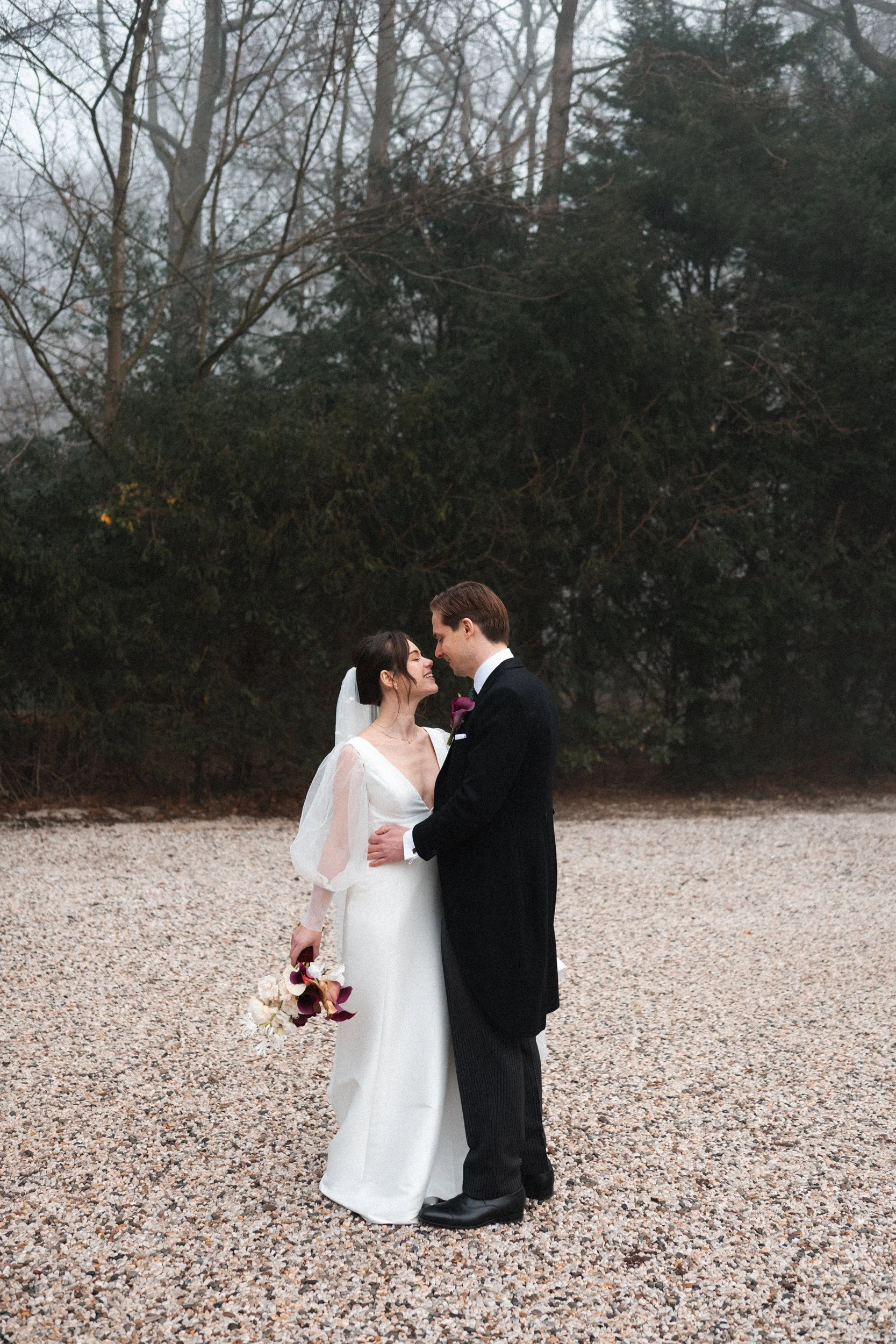 A bride and groom stand close together on a gravel area outdoors, gazing into each other's eyes, with trees in the background. The bride wears a white dress with sheer sleeves, holding a bouquet of dark and light flowers. The groom wears a black tuxe