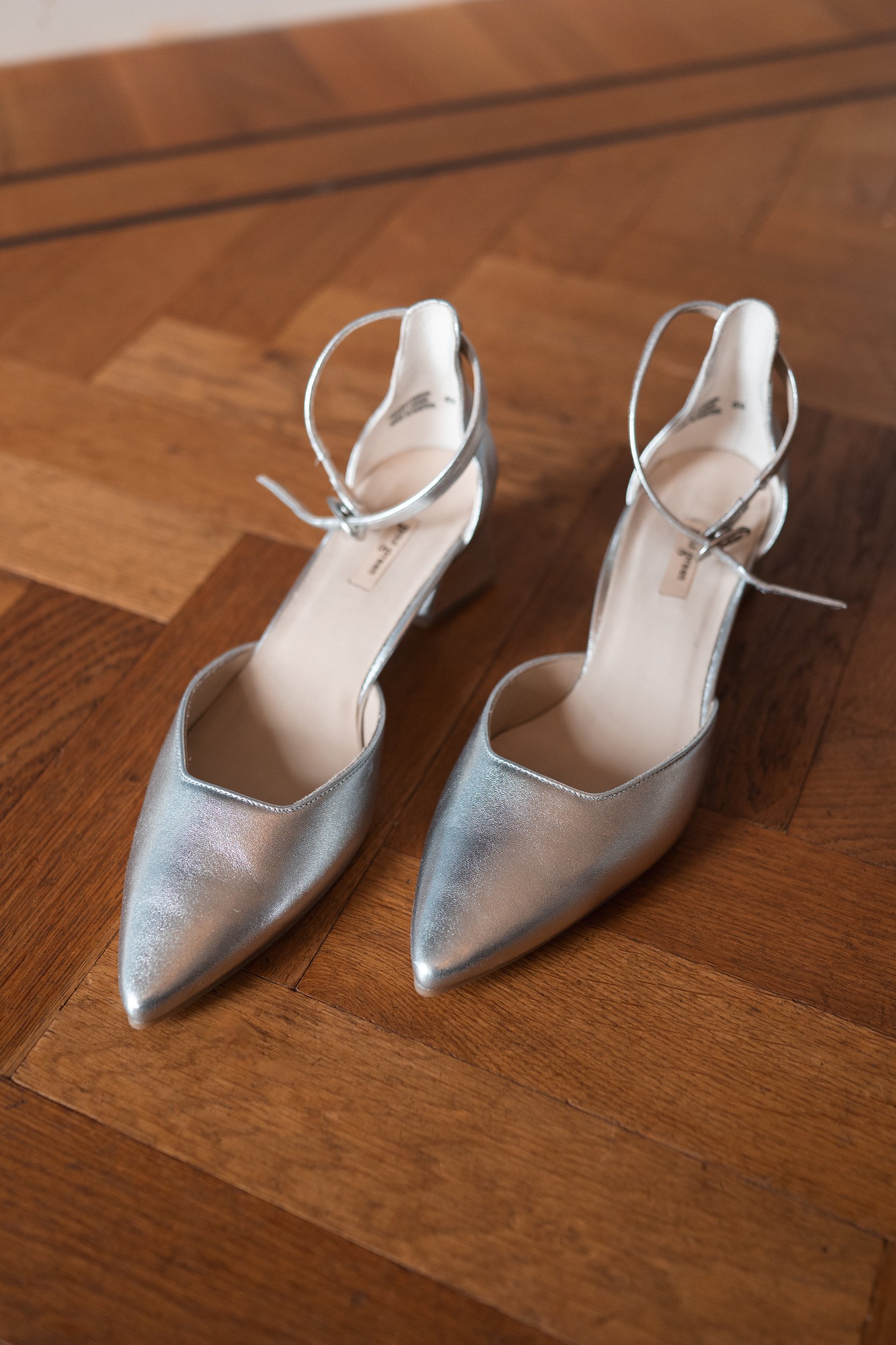 Silver pointed-toe high heels with ankle straps on wooden floor.