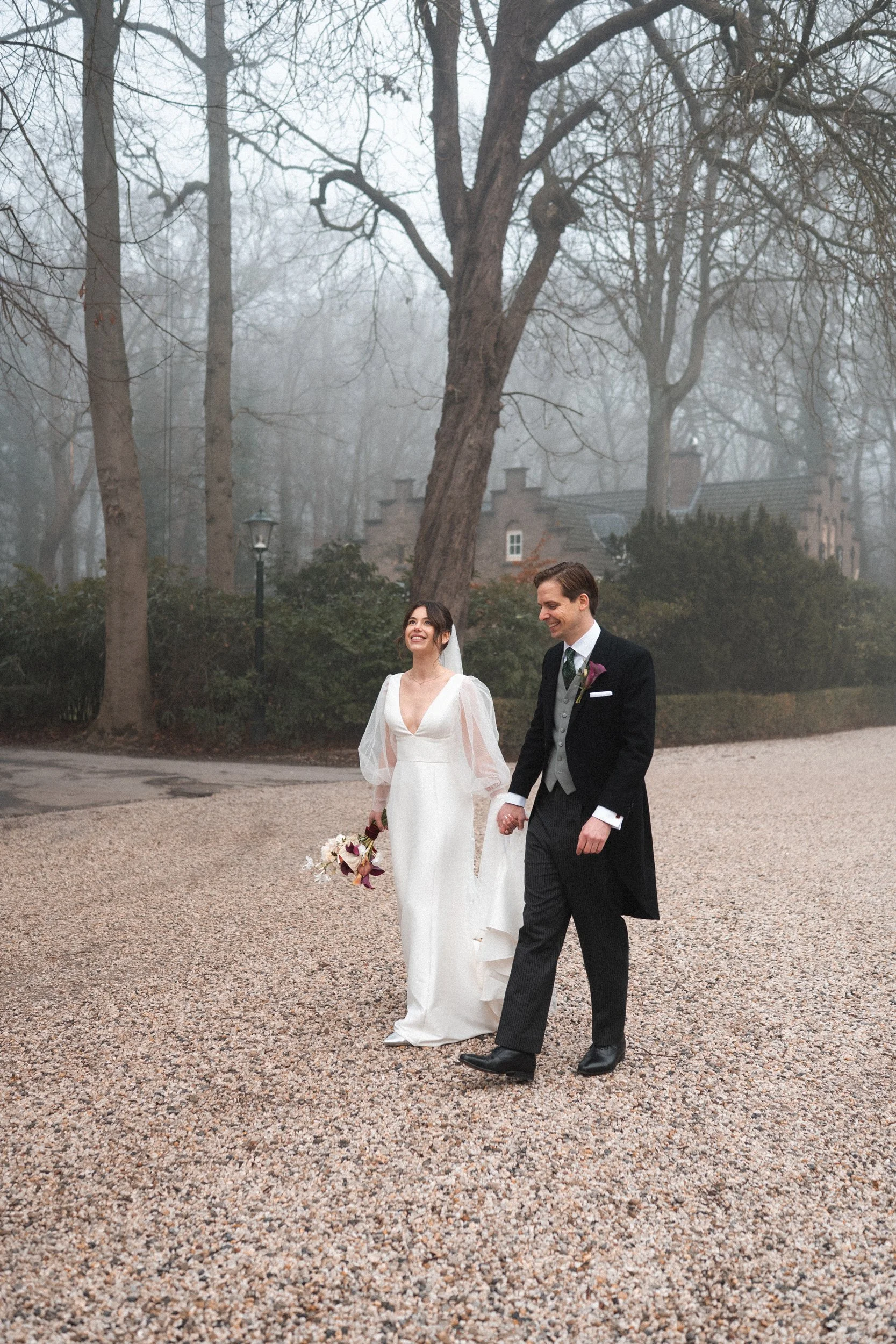 A bride and groom walking hand in hand outdoors on a foggy day, smiling and enjoying their wedding moment.