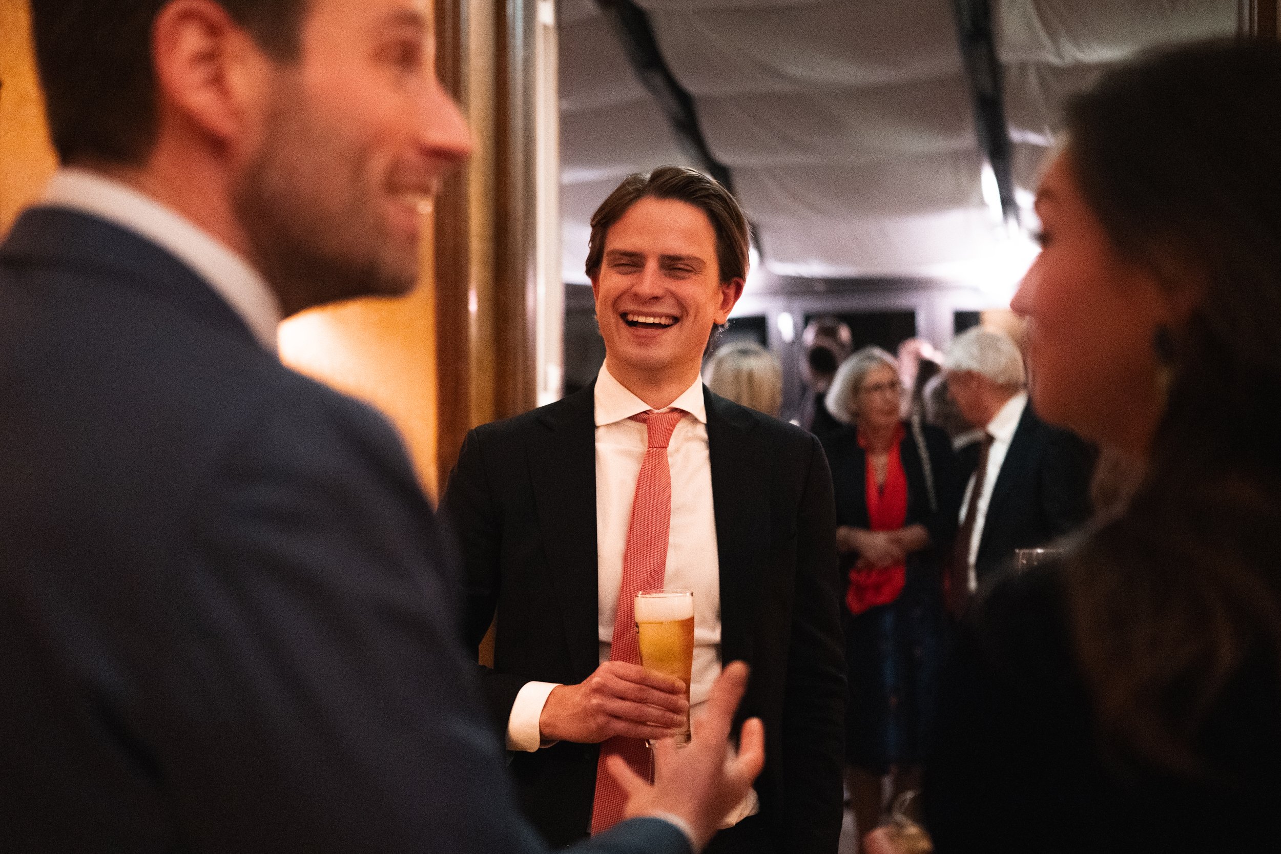 Man in a black suit holding a glass of beer, smiling at two people in conversation at a social gathering.