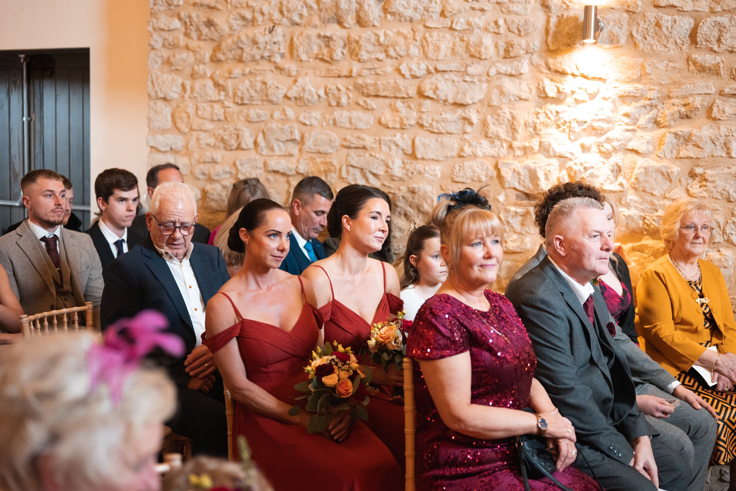 Guests seated at a wedding ceremony or event in a room with a stone wall, predominantly dressed in formal attire, with some women holding bouquets of flowers.