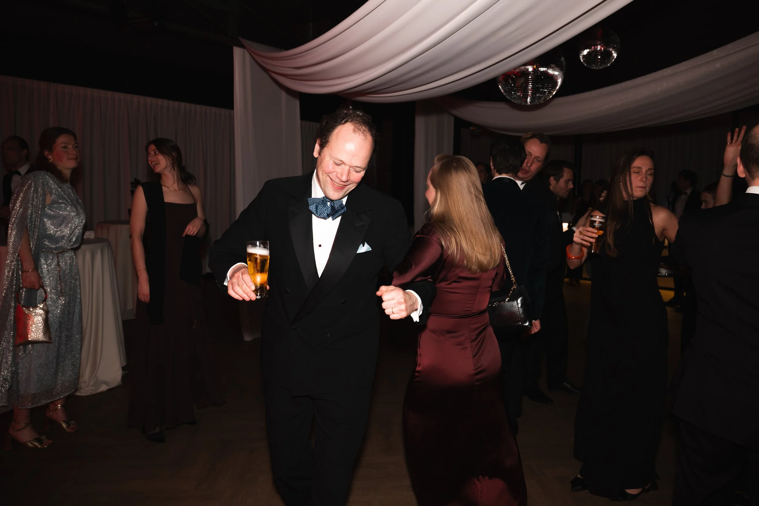 People dancing at a formal event or party, with men in tuxedos and women in evening dresses, some holding drinks