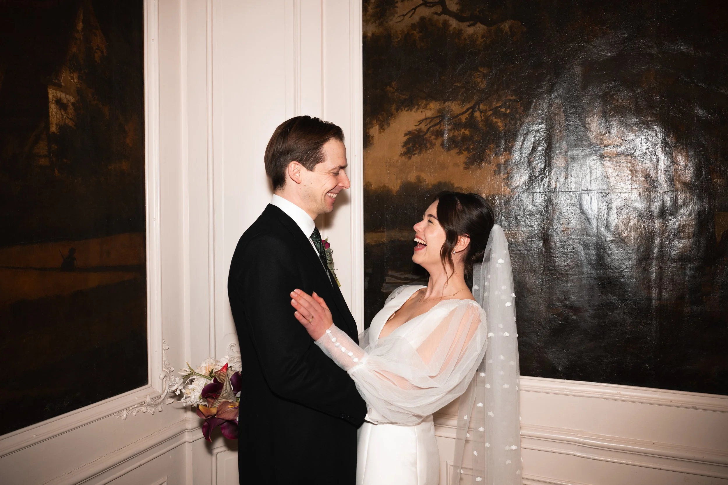 A bride and groom stand facing each other indoors, smiling and laughing, with a dark large painting or mural in the background.