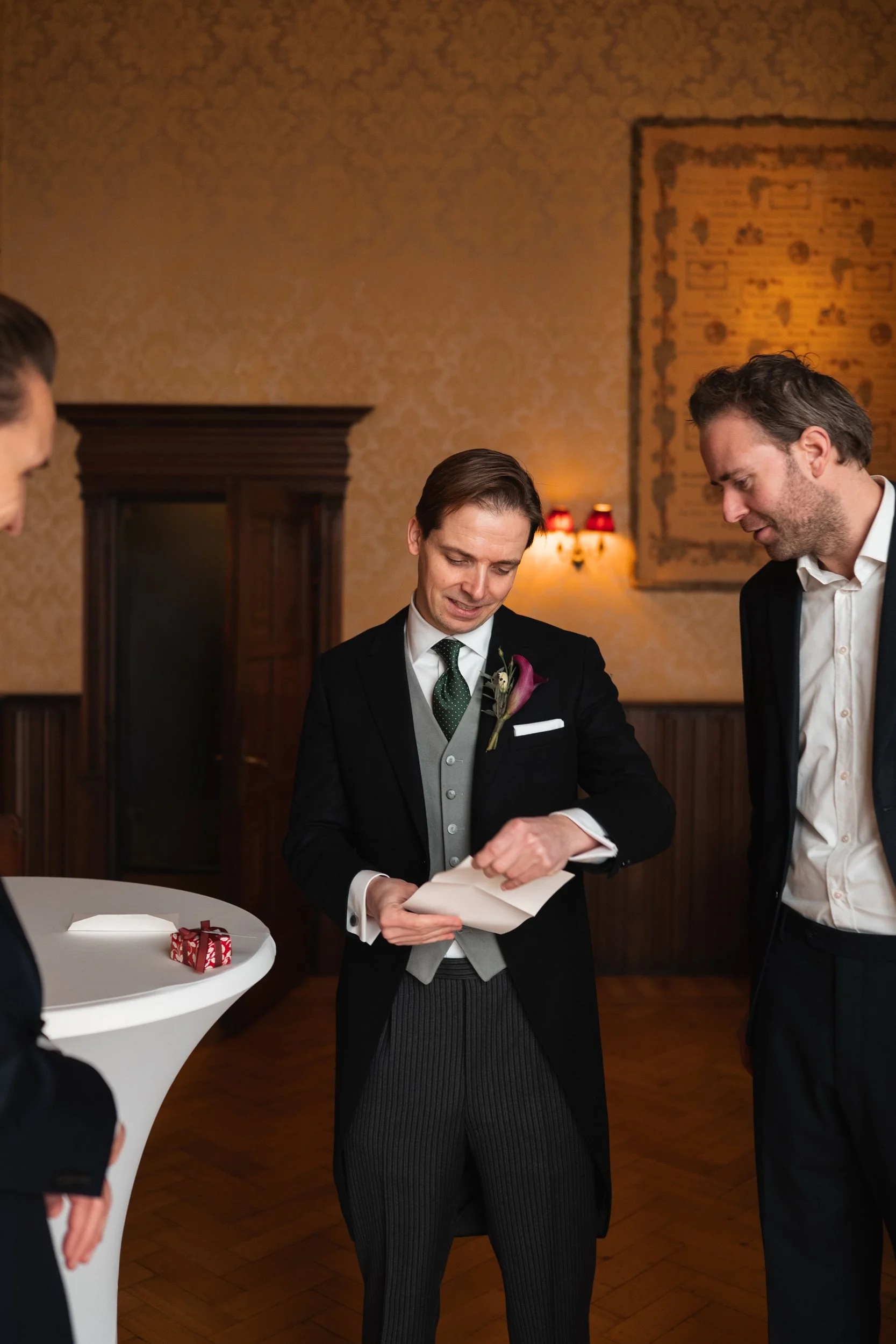 A groom reading a heartfelt note or speech during a wedding reception, surrounded by friends, in a warmly decorated room with vintage wallpaper and soft lighting.