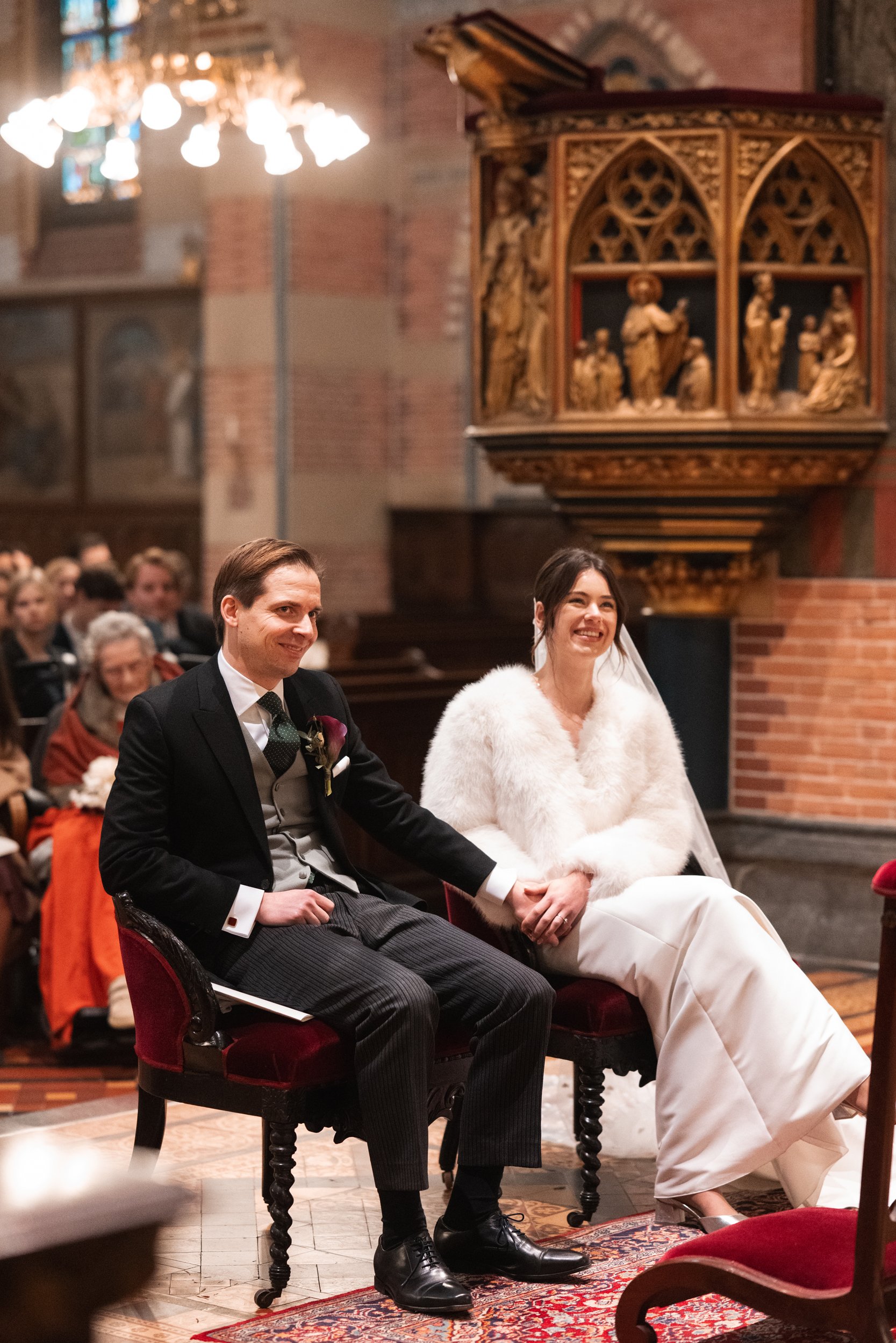 A bride and groom sitting on chairs inside a church during their wedding ceremony, holding hands and smiling.