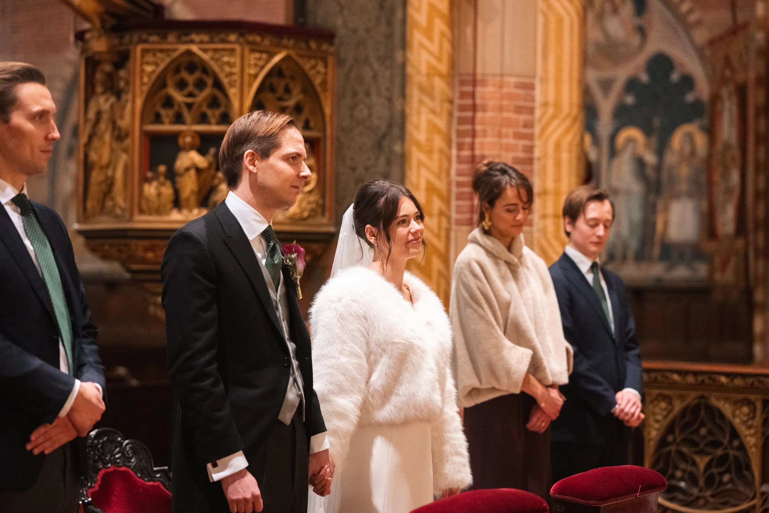 A bride and groom stand with three other individuals during a wedding ceremony inside a church with ornate decorations and artwork in the background.