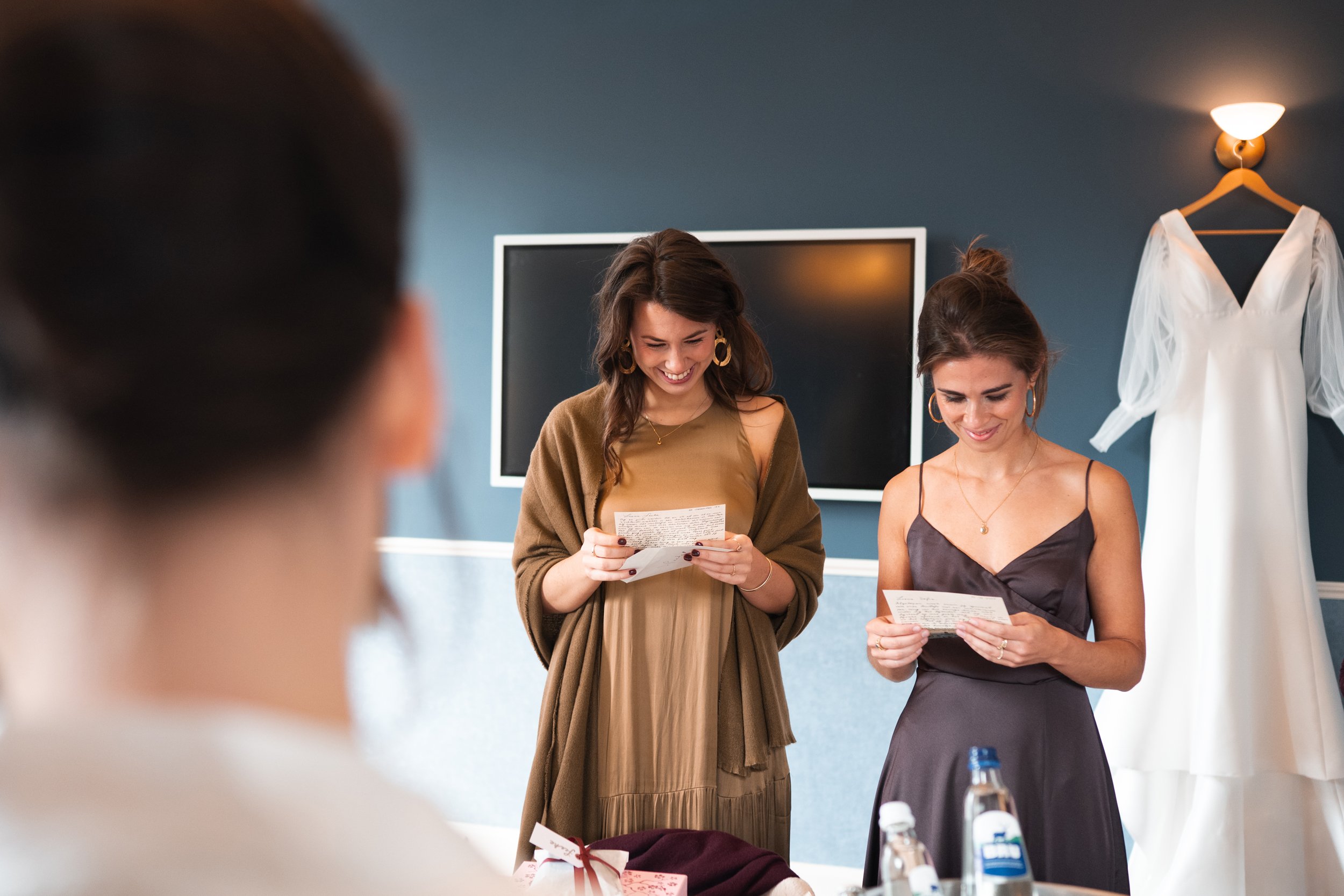 Two women in dresses reading notes in a room with a large white dress hanging on the wall
