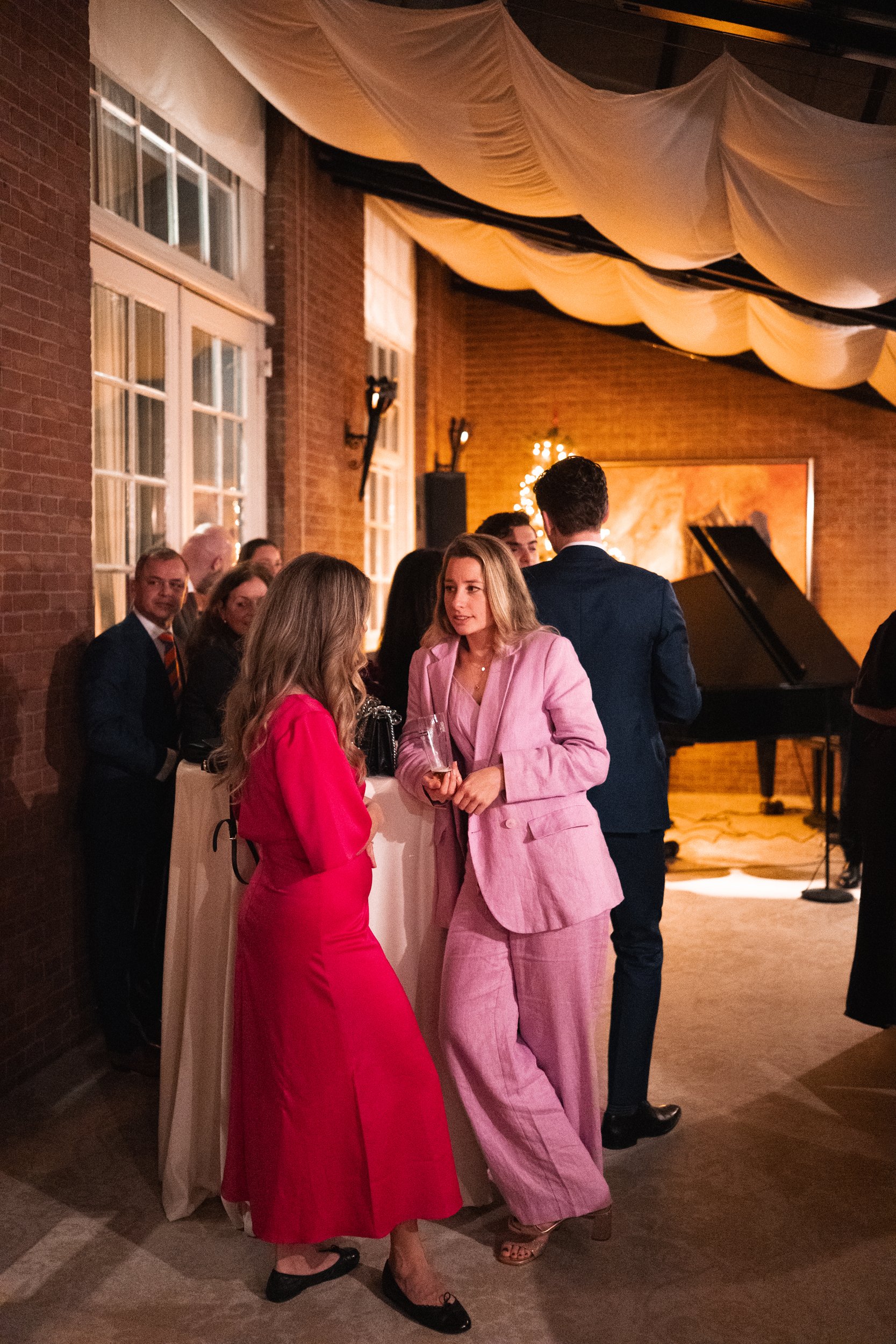 People at an indoor social event, including women in pink and red dresses, talking near a high table, with a grand piano in the background and warm indoor lighting.