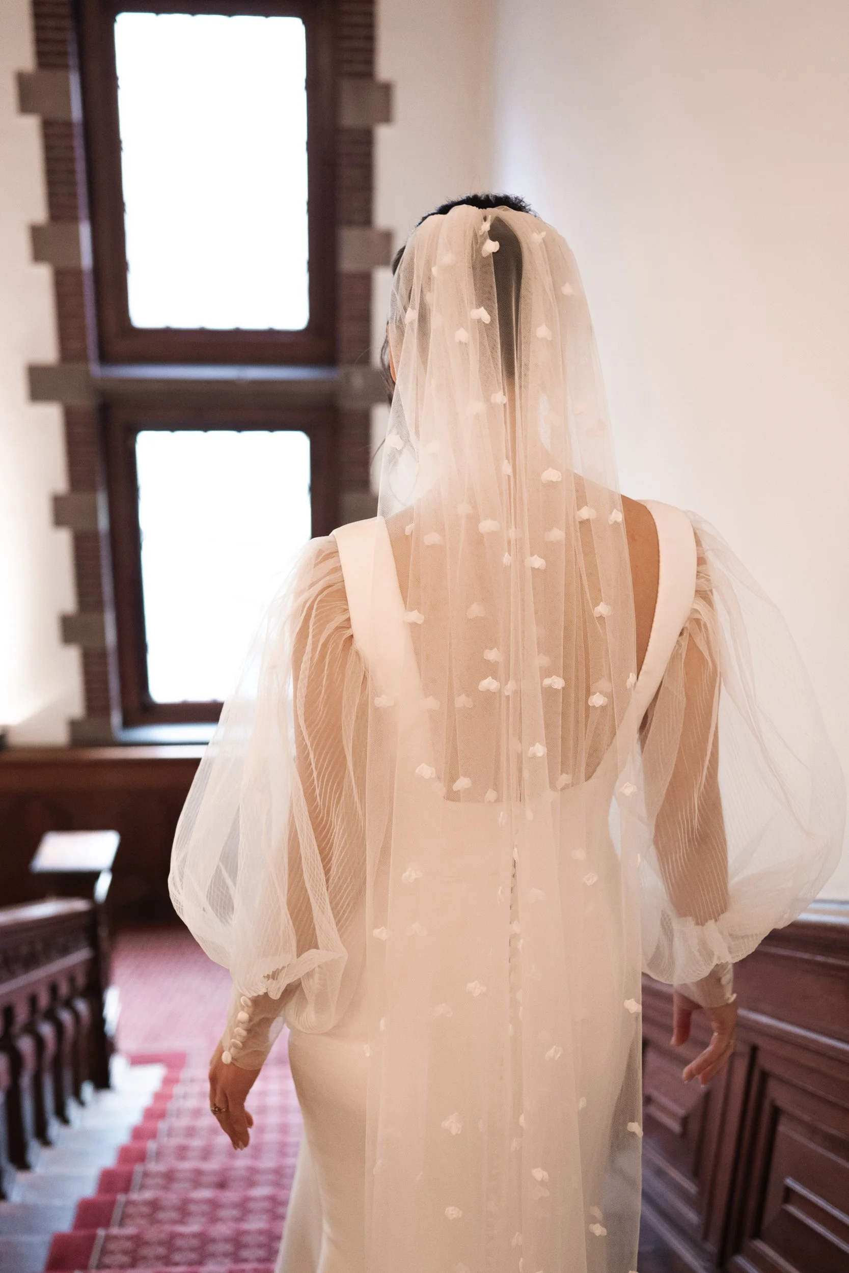 Back view of a woman in a wedding dress with a veil, standing in a wood-paneled room with stained glass windows.
