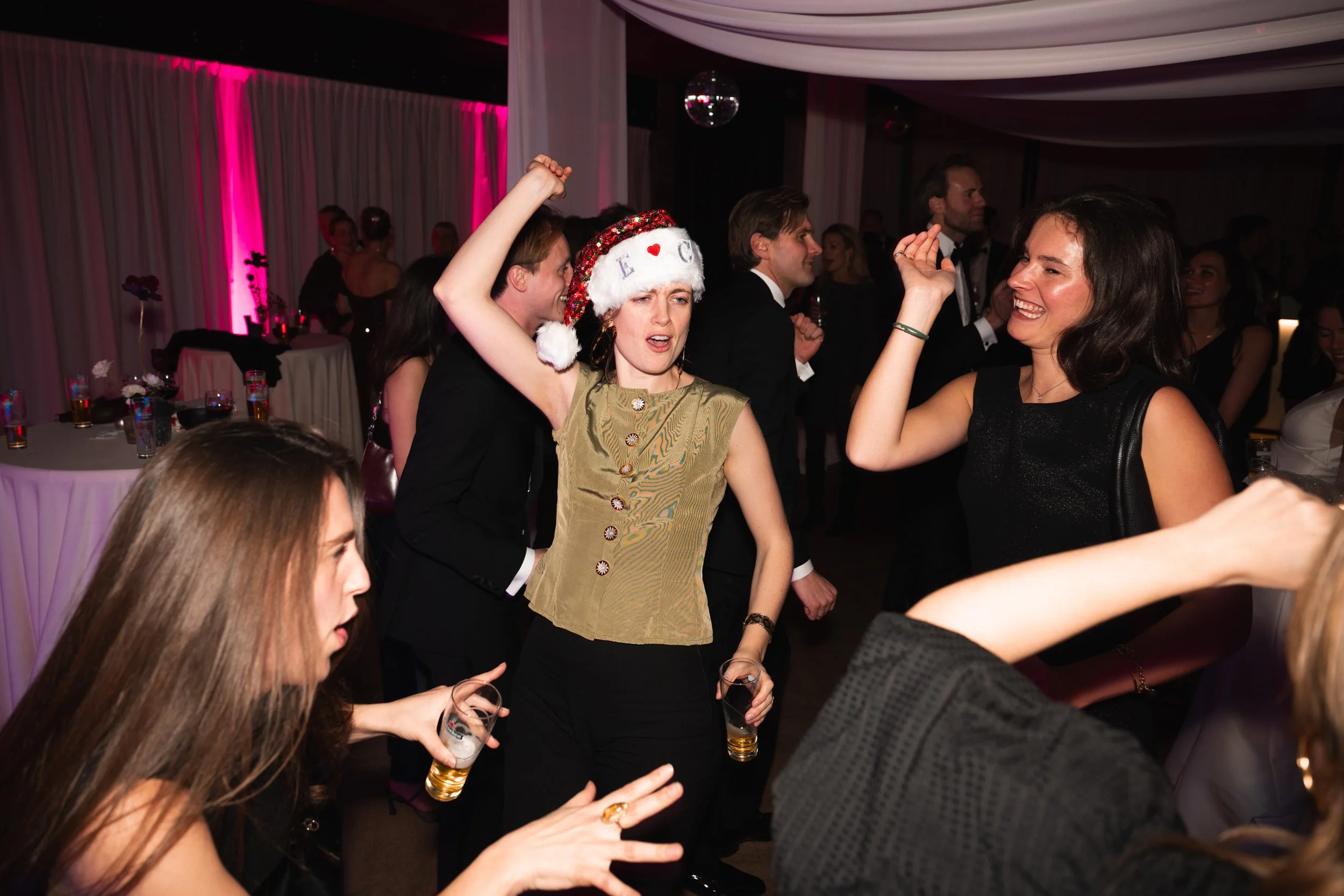 Group of people dancing at a festive party with a woman wearing a holiday hat and others holding drinks, smiling and enjoying.