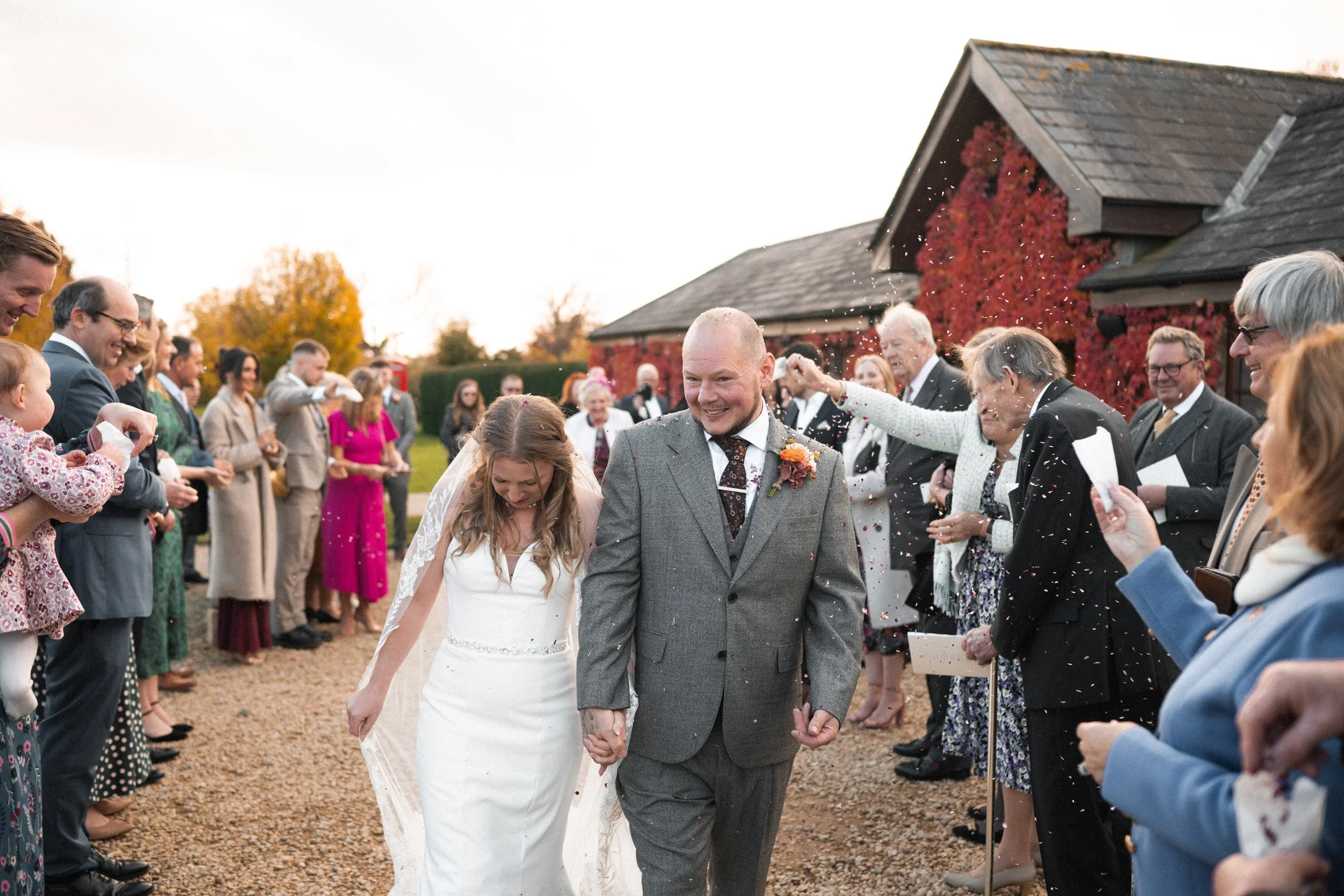 Bride and groom walking hand-in-hand through a crowd of smiling wedding guests outside on a gravel path, with guests throwing confetti in celebration.