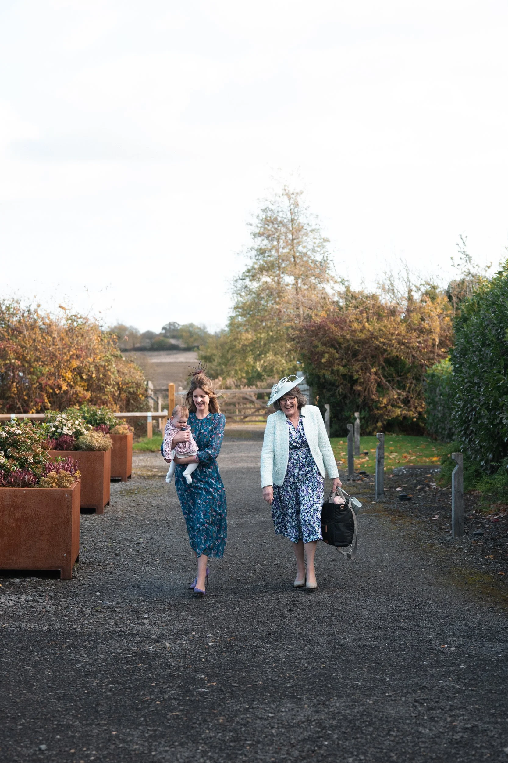 Two women walking outdoors on a gravel path, one carrying a baby, surrounded by autumn scenery with trees and bushes.