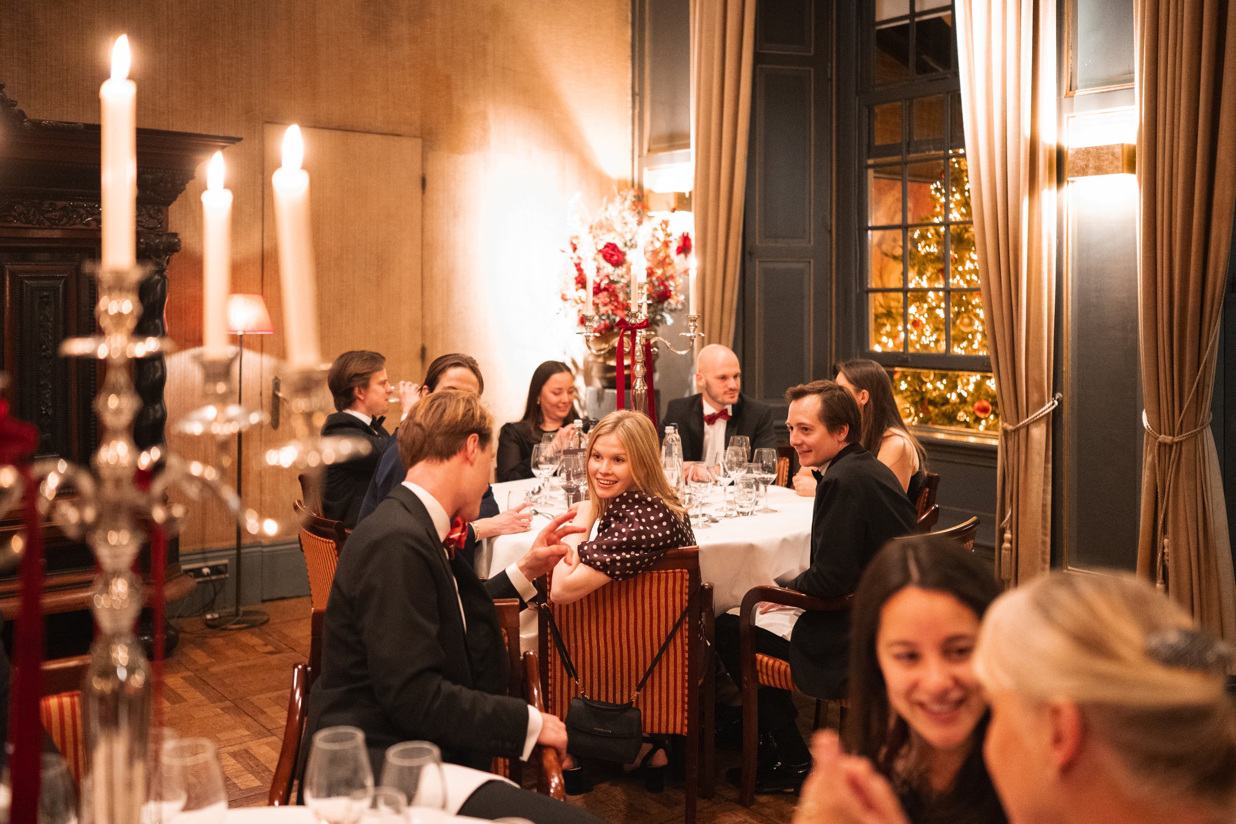 People dressed in formal attire sit around a dining table in a decorated festive room, with a Christmas tree seen through the window.