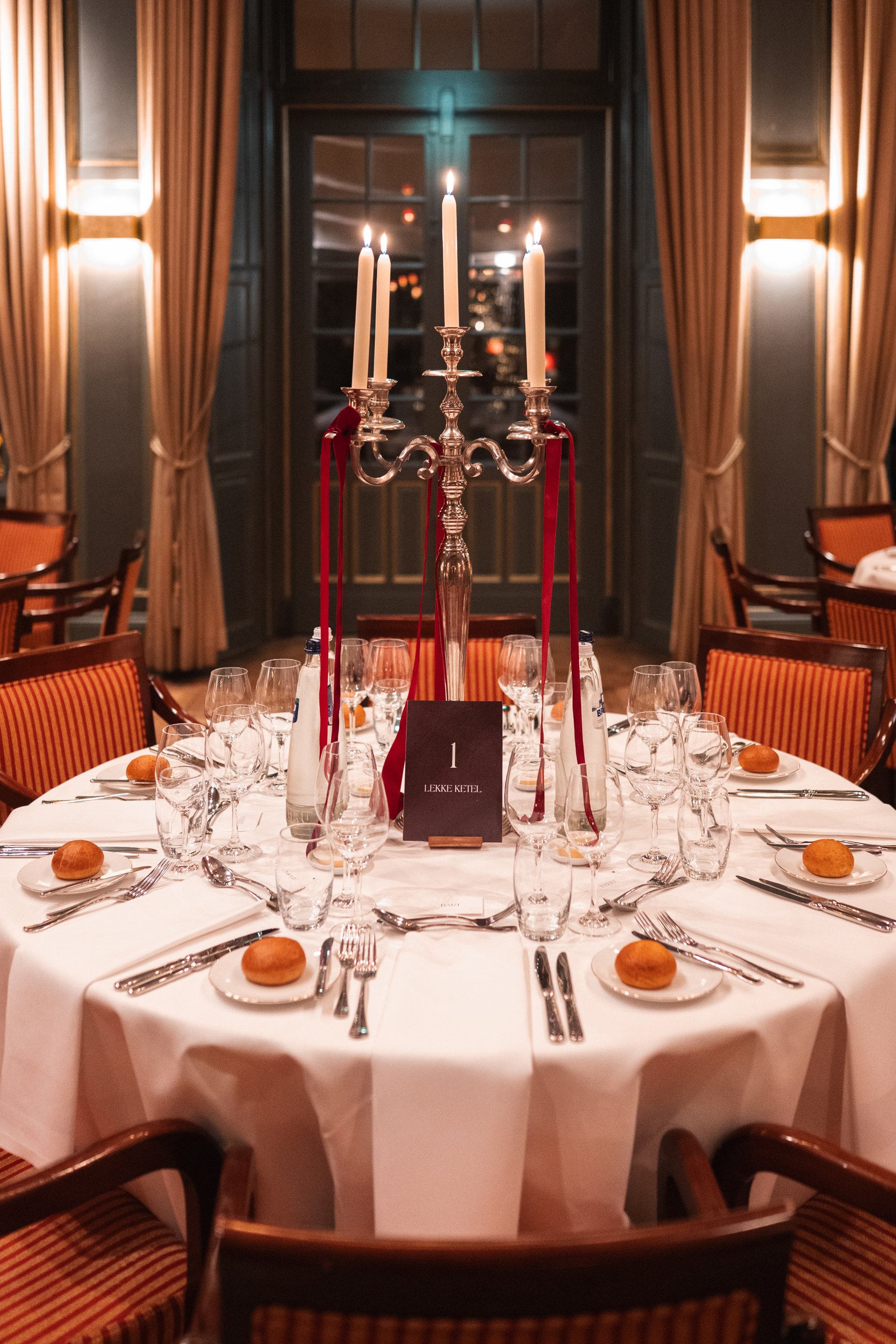 Elegant round dinner table set with a white tablecloth, multiple glasses, silverware, small bread rolls on plates, and a silver candelabrum with lit candles at the center in a sophisticated dining room with warm lighting and draped curtains.