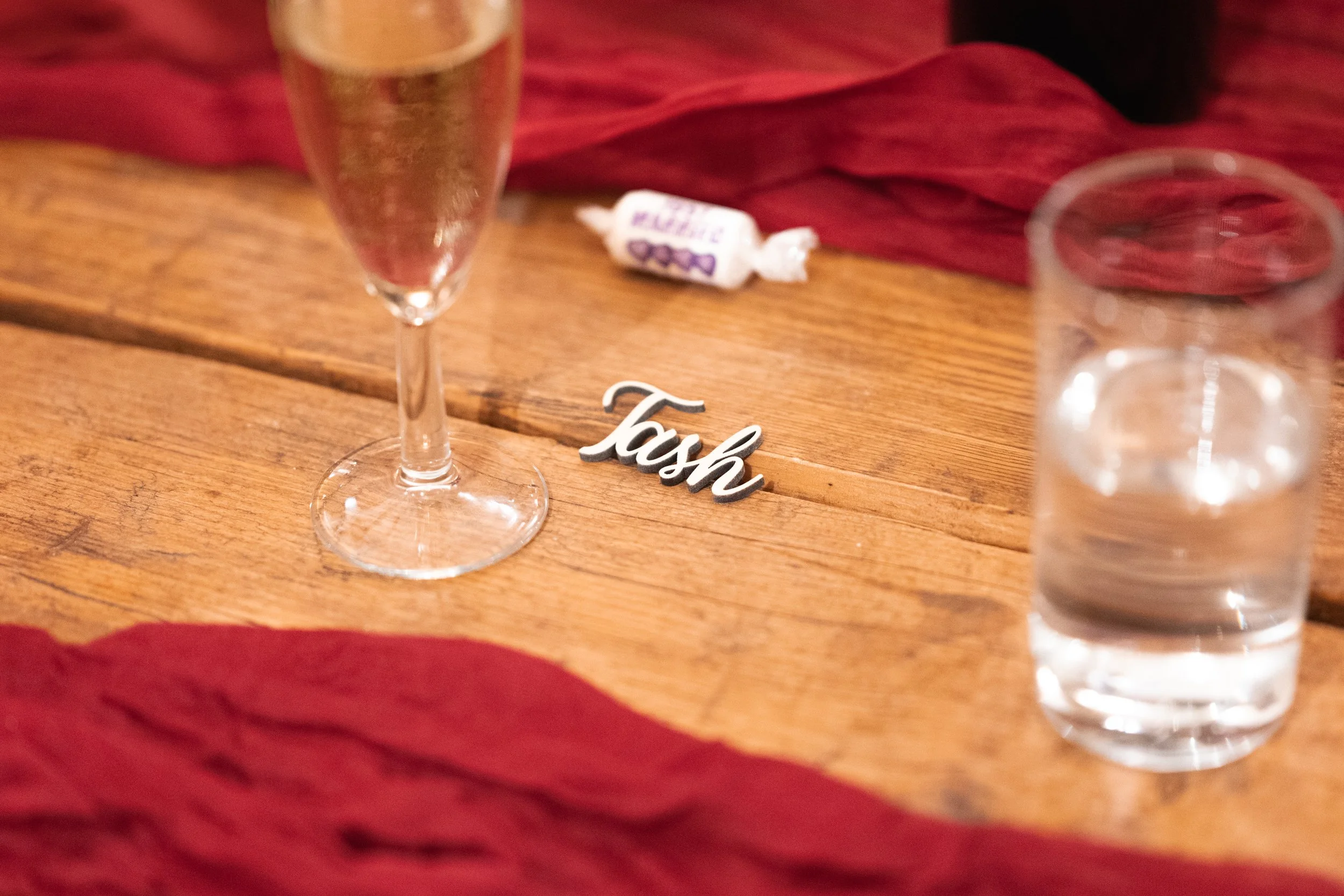 A wooden table with a glass of champagne, a glass of water, a small piece of paper reading 'Tash,' and a wrapped candy. Red cloth is draped along the edges of the table.