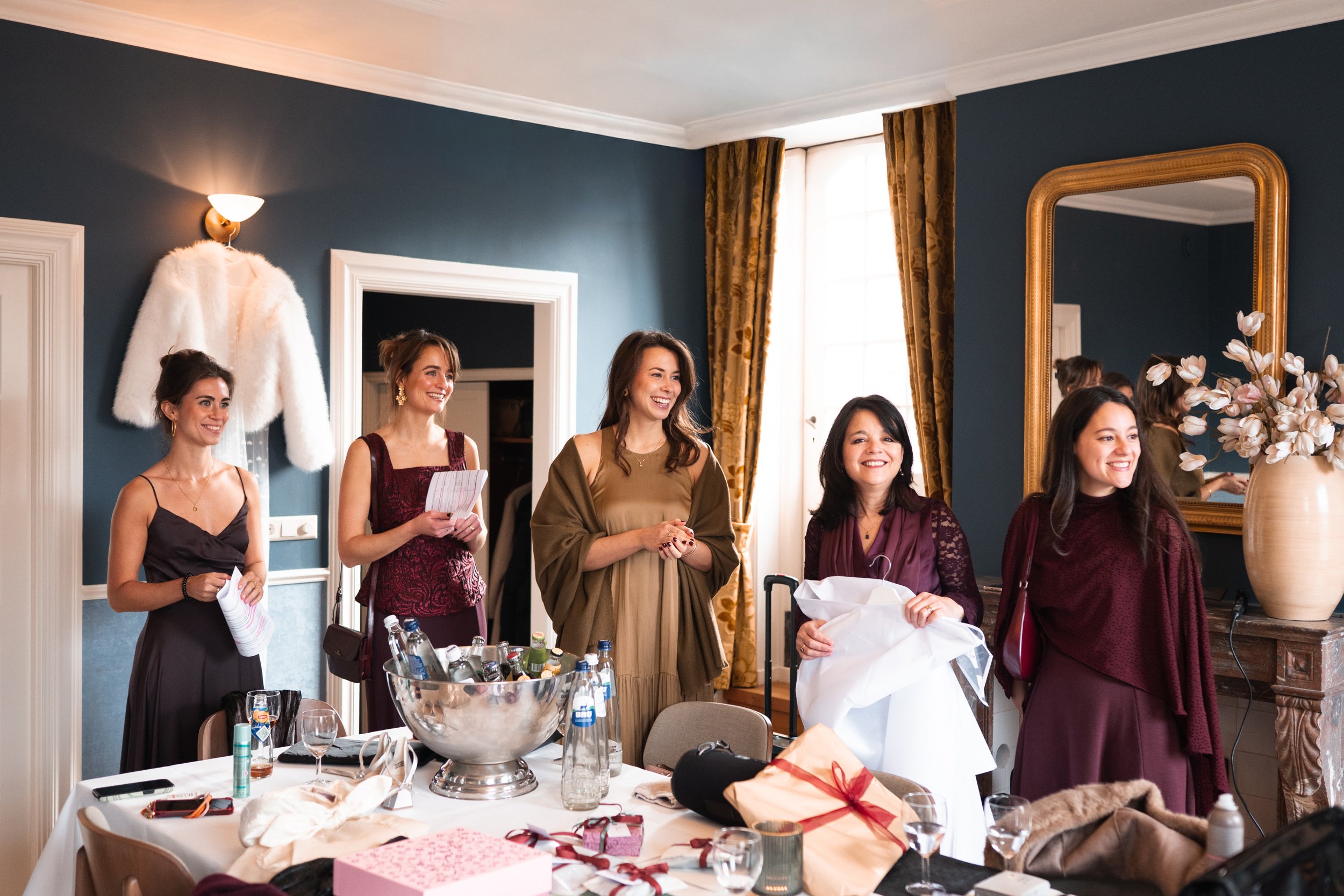 Group of women smiling and standing in a decorated room, with gifts and drinks on the table in front of them, preparing for a celebration or gathering.
